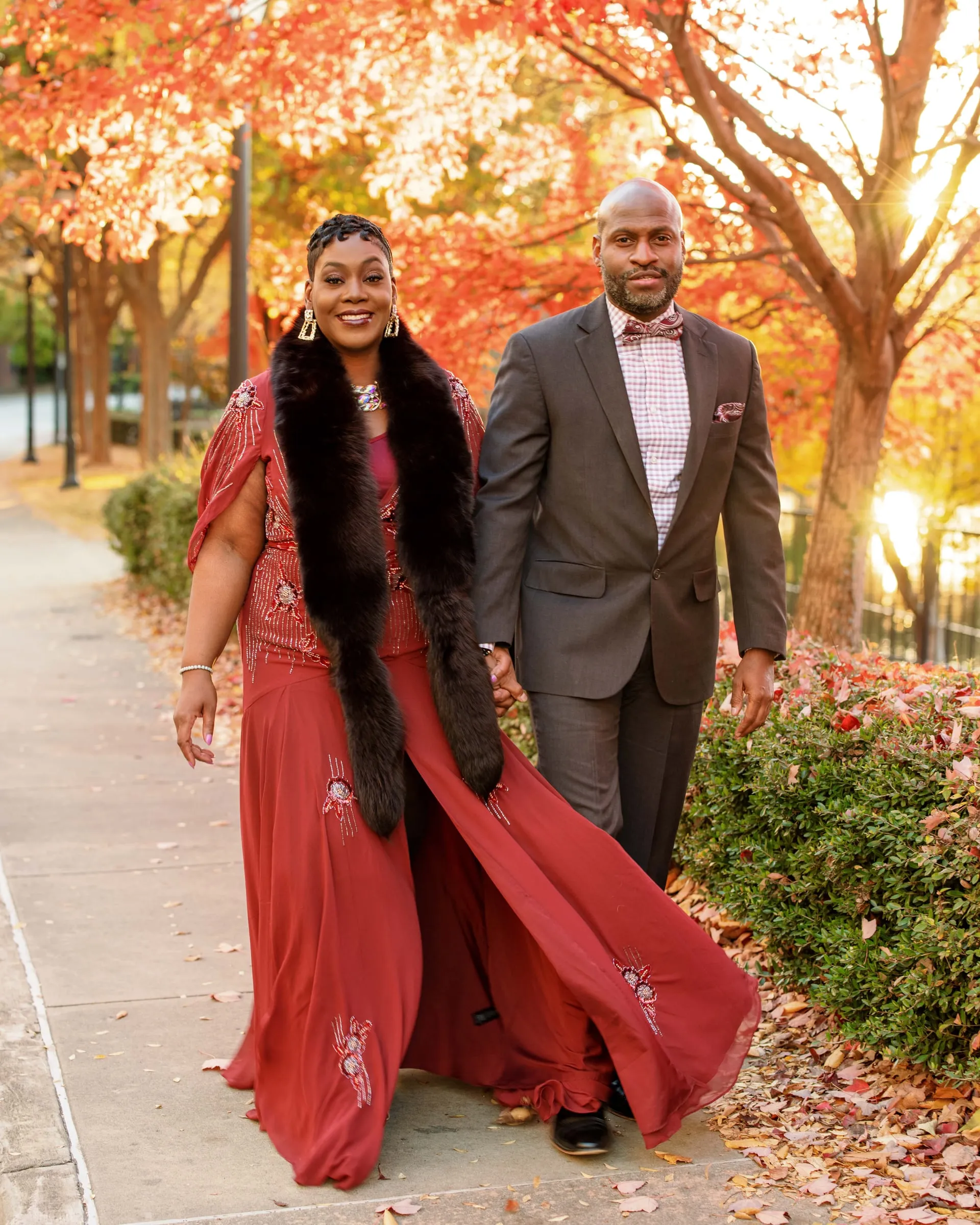 A couple strolling a lakeside path under autumn leaves, she in red and a fur stole.