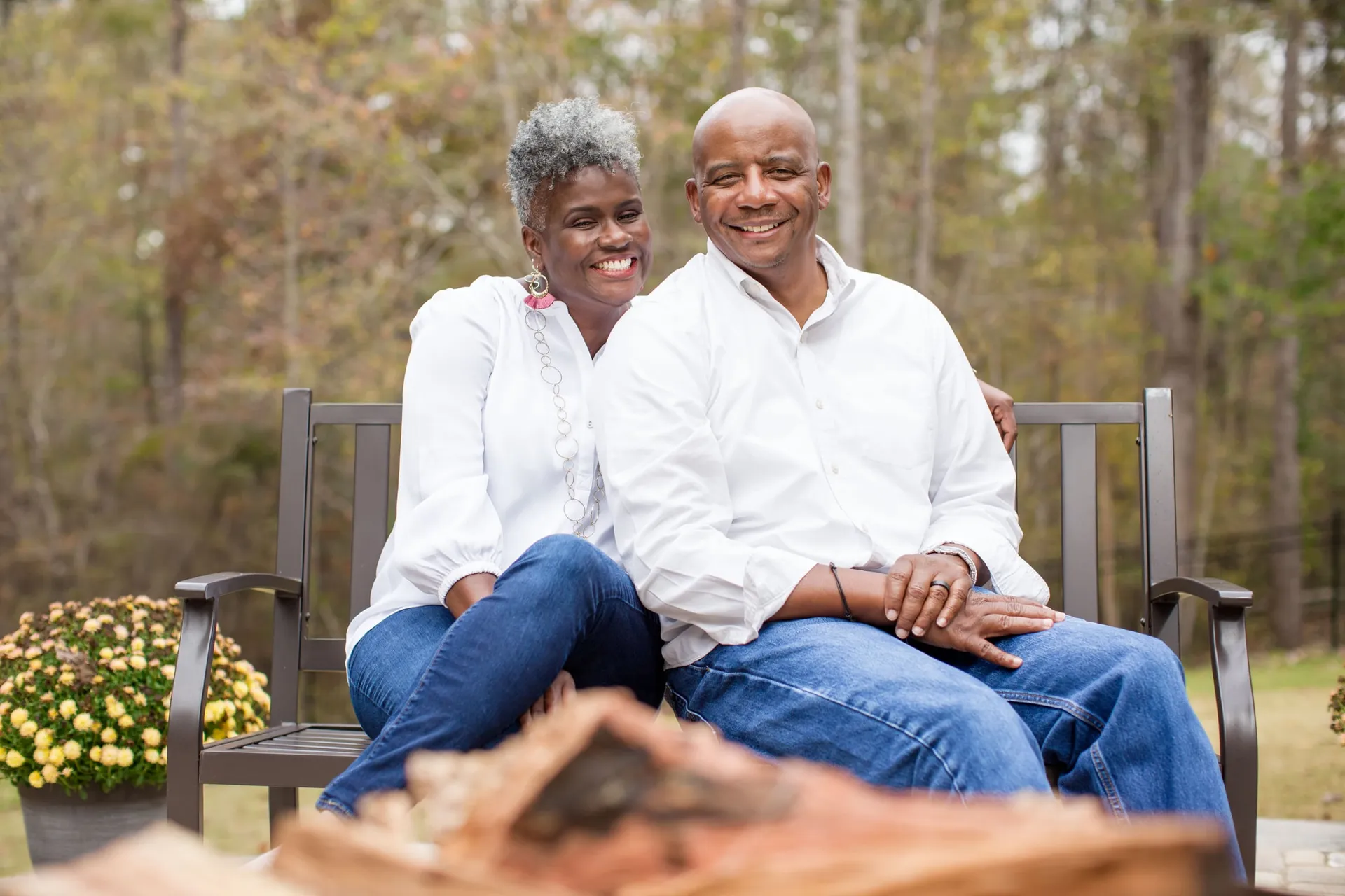A couple in white shirts on a bench among pines beside a pot of yellow mums.