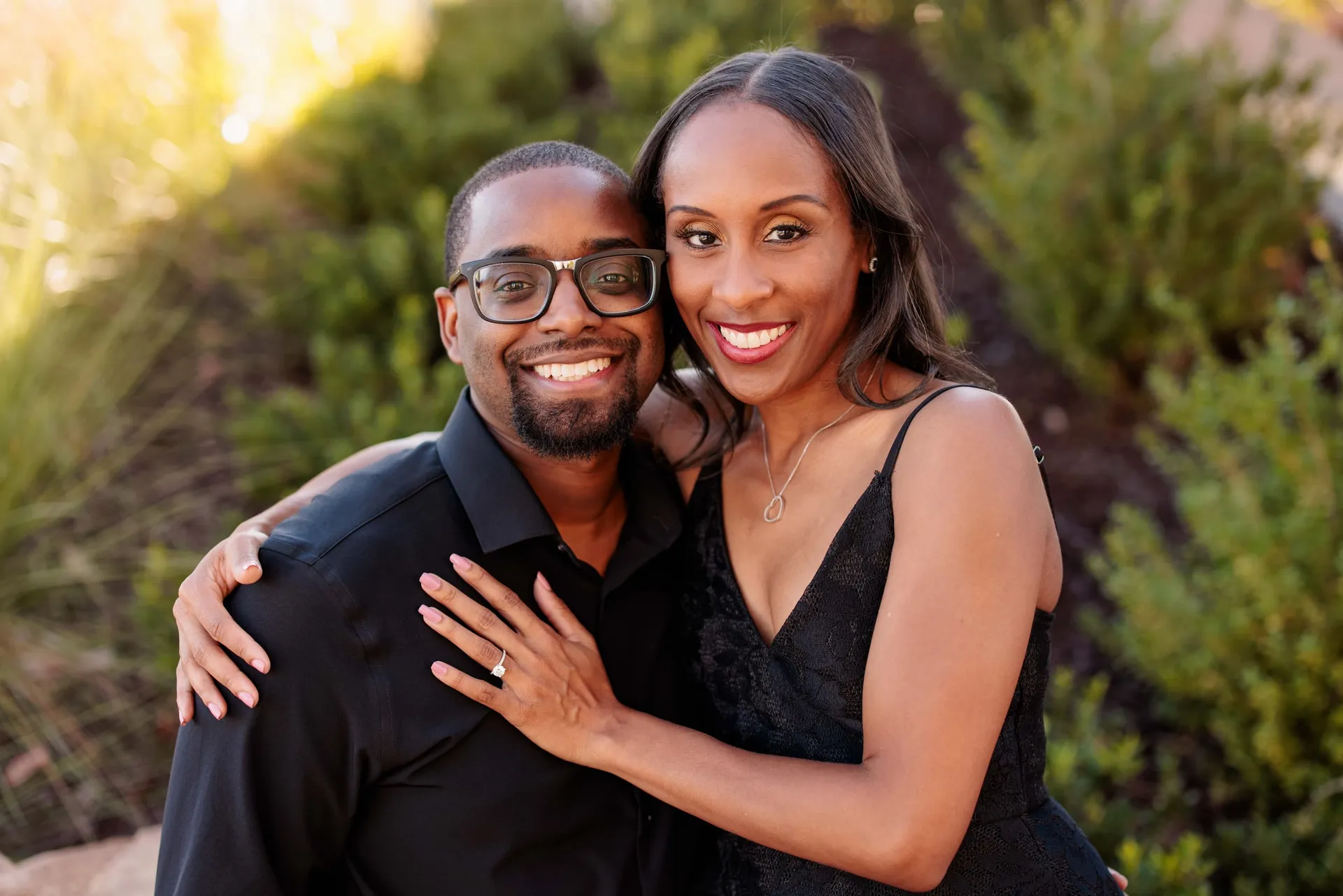 An engaged couple in black smiling at the camera, her ring hand resting on his chest.
