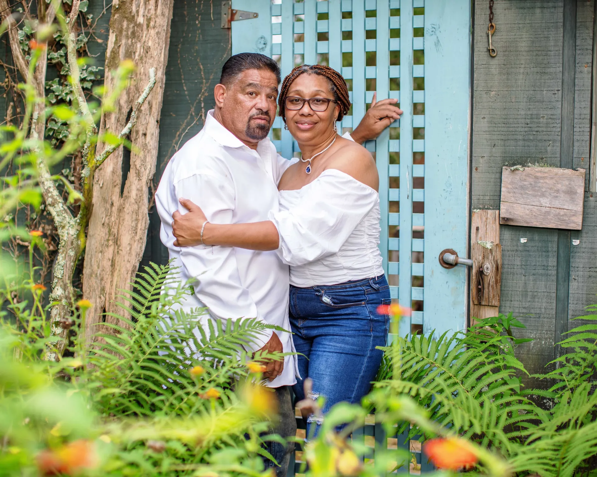 An engaged couple leaning against a weathered teal garden door among ferns and wildflowers.