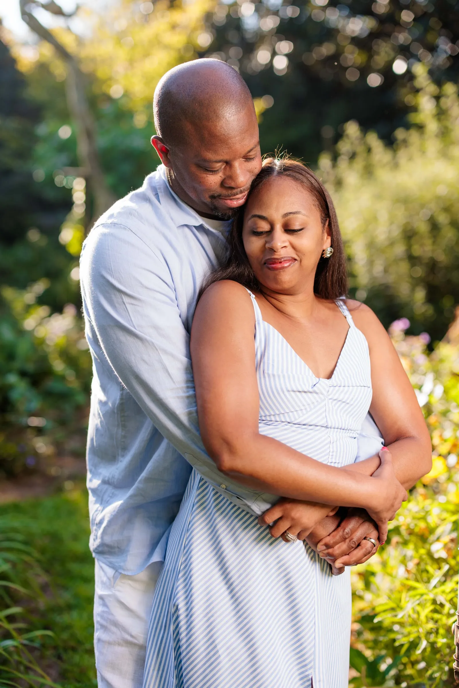 A man in a light blue shirt holding his partner from behind in a sunlit garden.