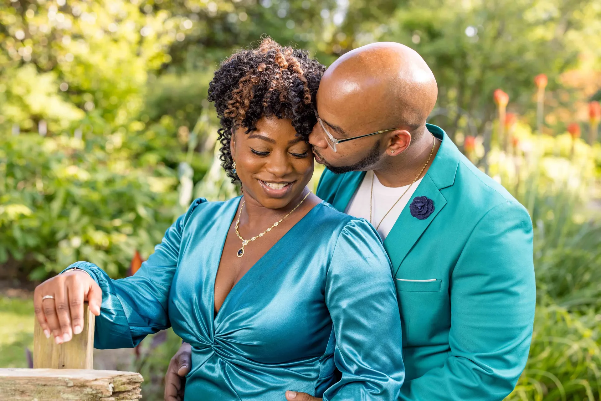A man in a teal jacket kissing his partner's temple in a sunlit garden.