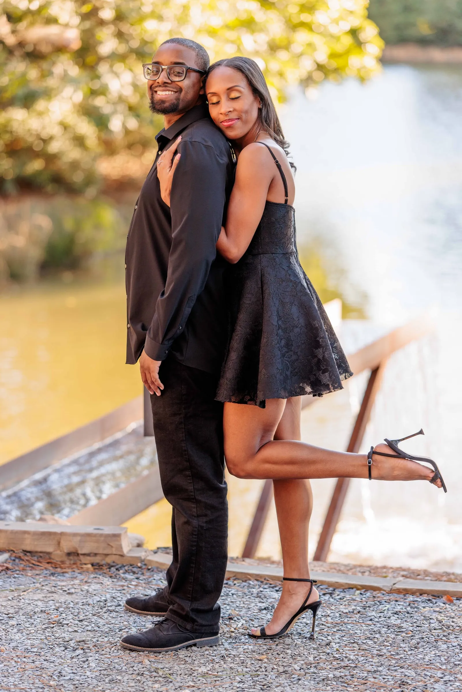An engaged couple on a lakeside footbridge, she hugging him from behind, one heel kicked up.