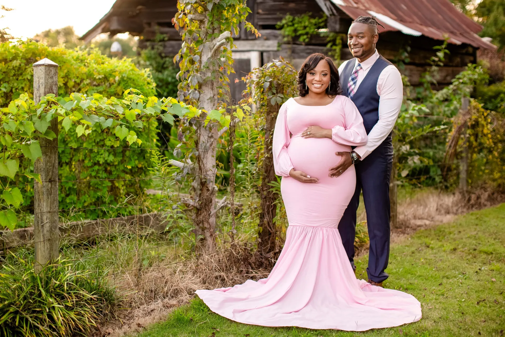 An expectant mother in a pink gown with her partner beside grapevines and a rustic cabin.