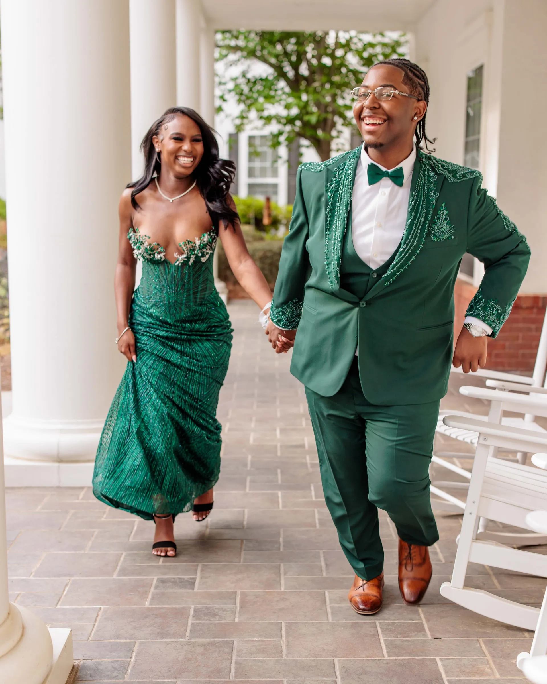 A prom couple in matching emerald green walking hand in hand along a columned porch.