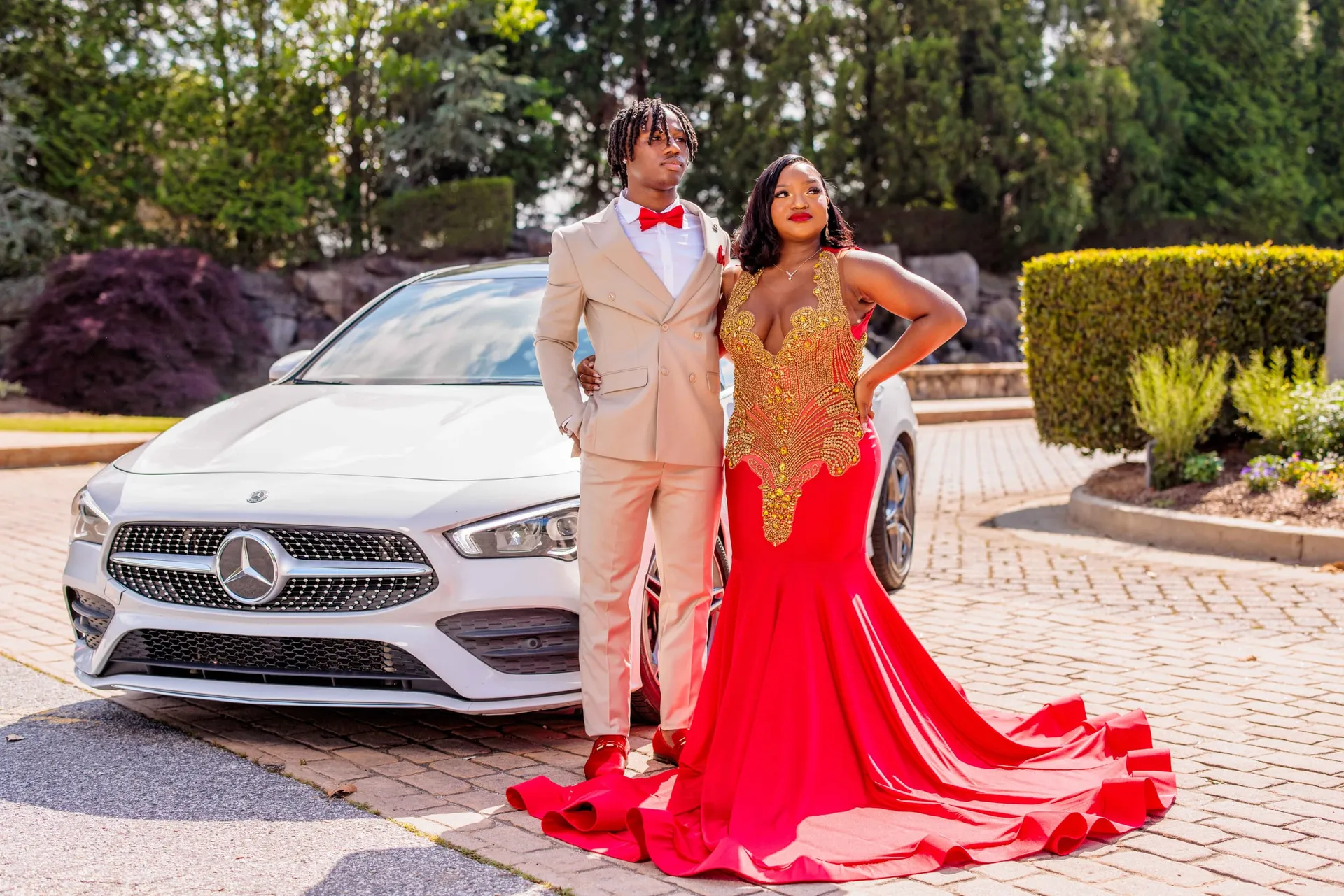 A prom couple in a red gown and tan suit posing beside a white Mercedes.