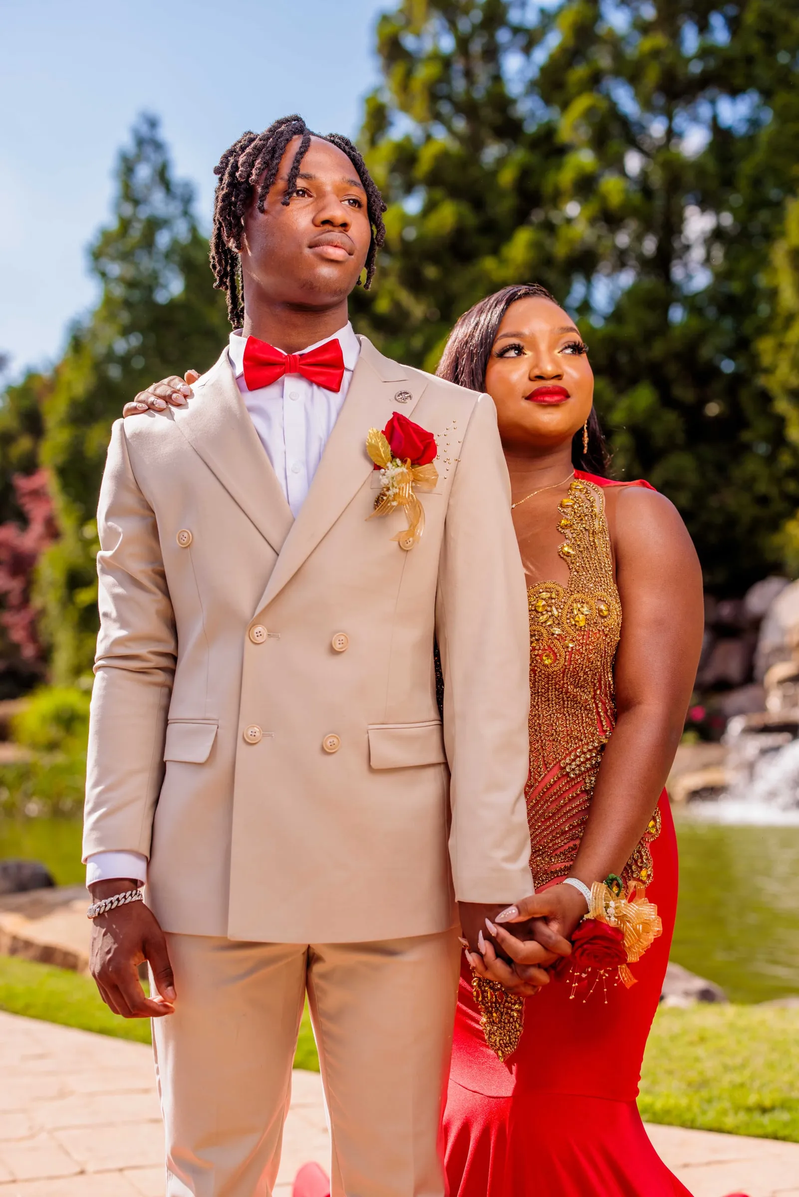 A prom couple side by side in a tan suit and red beaded gown beneath pines.