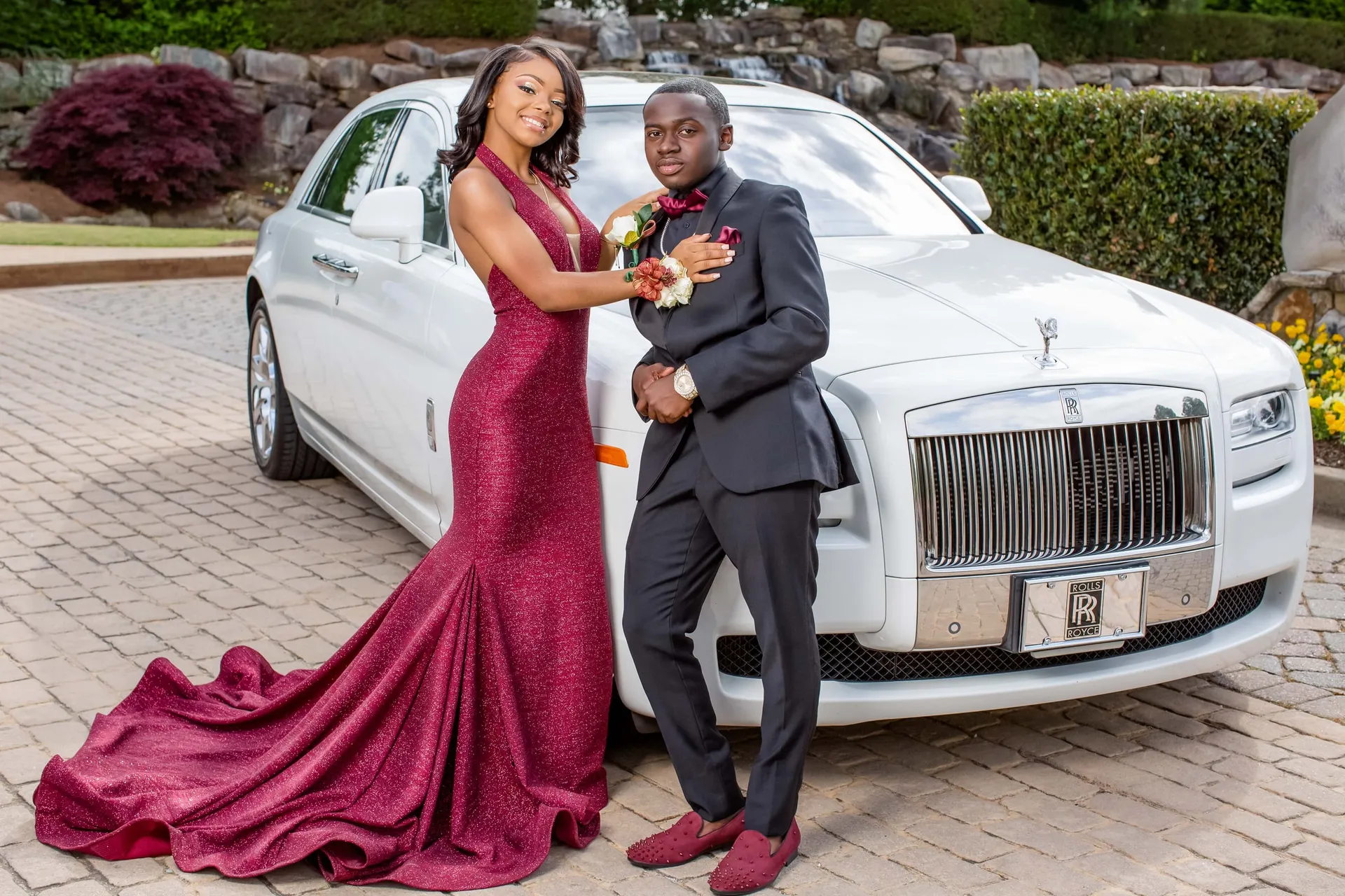 A prom couple in burgundy and black posing beside a white Rolls-Royce at a rock garden.