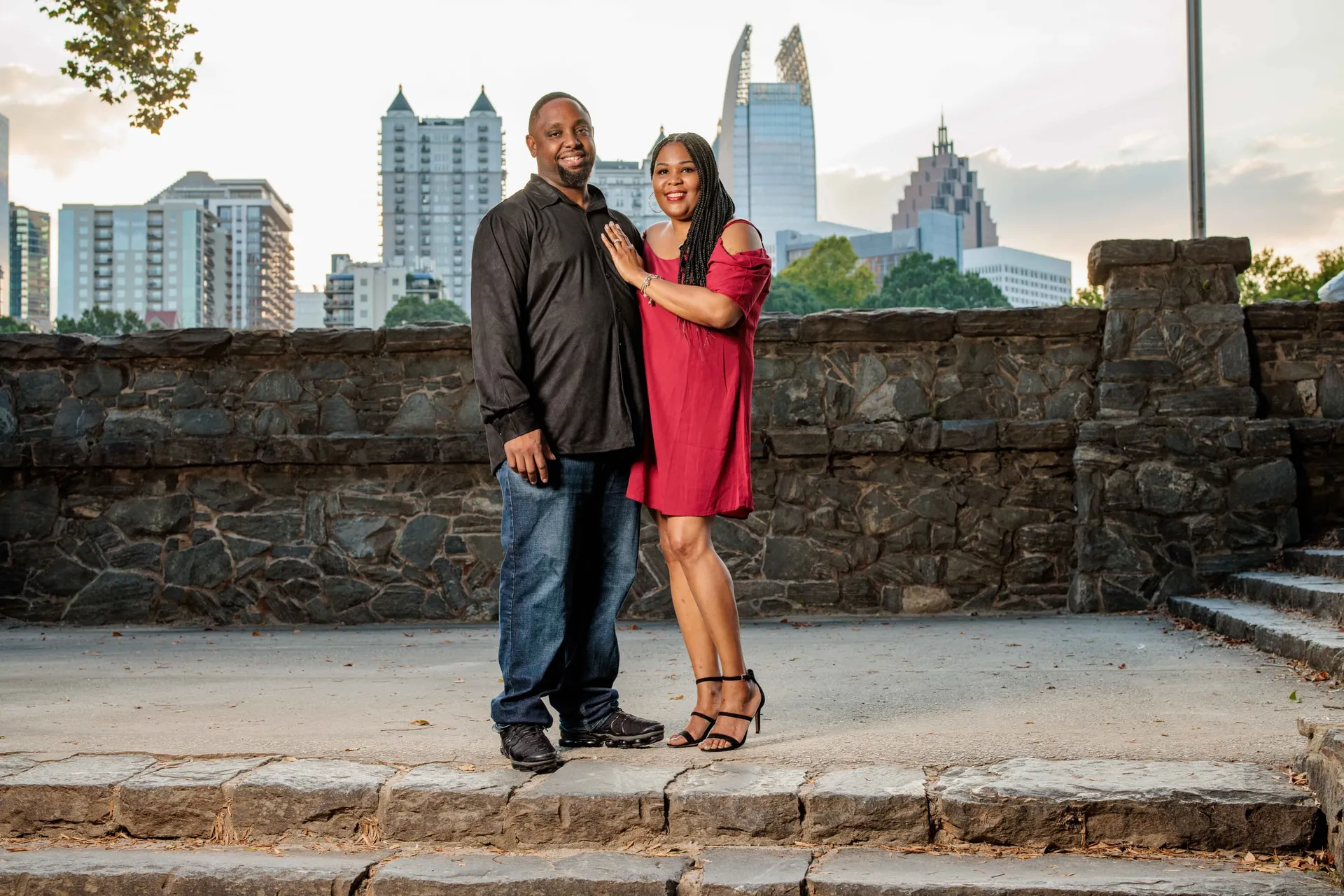 A couple on the stone wall at Piedmont Park with the Atlanta skyline rising behind them.