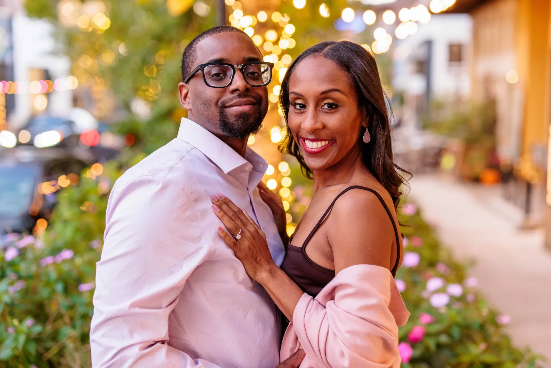 An engaged couple close together under warm string lights on a town street at dusk.