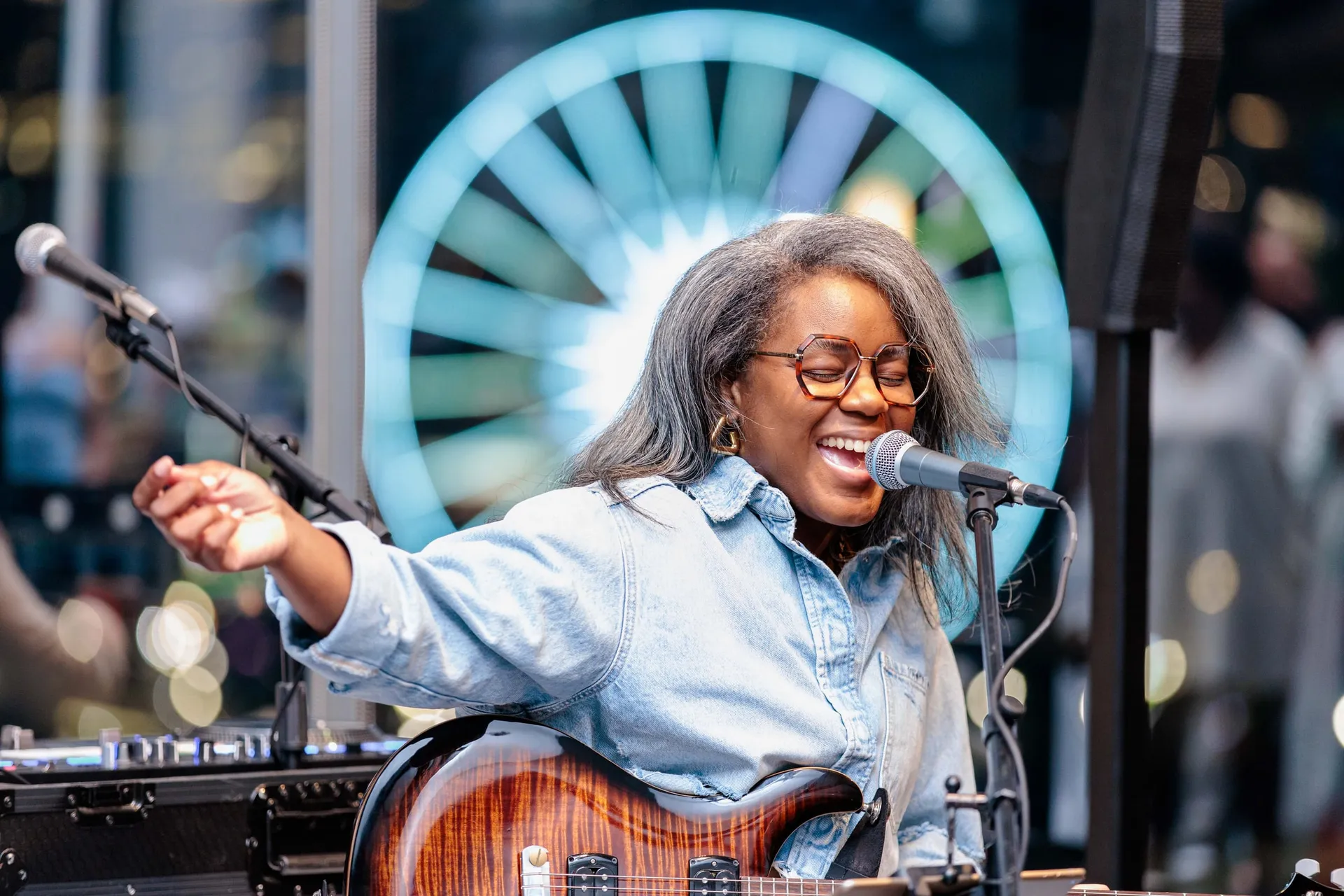 A singer with a guitar laughing into the microphone, a lit Ferris wheel glowing behind her.