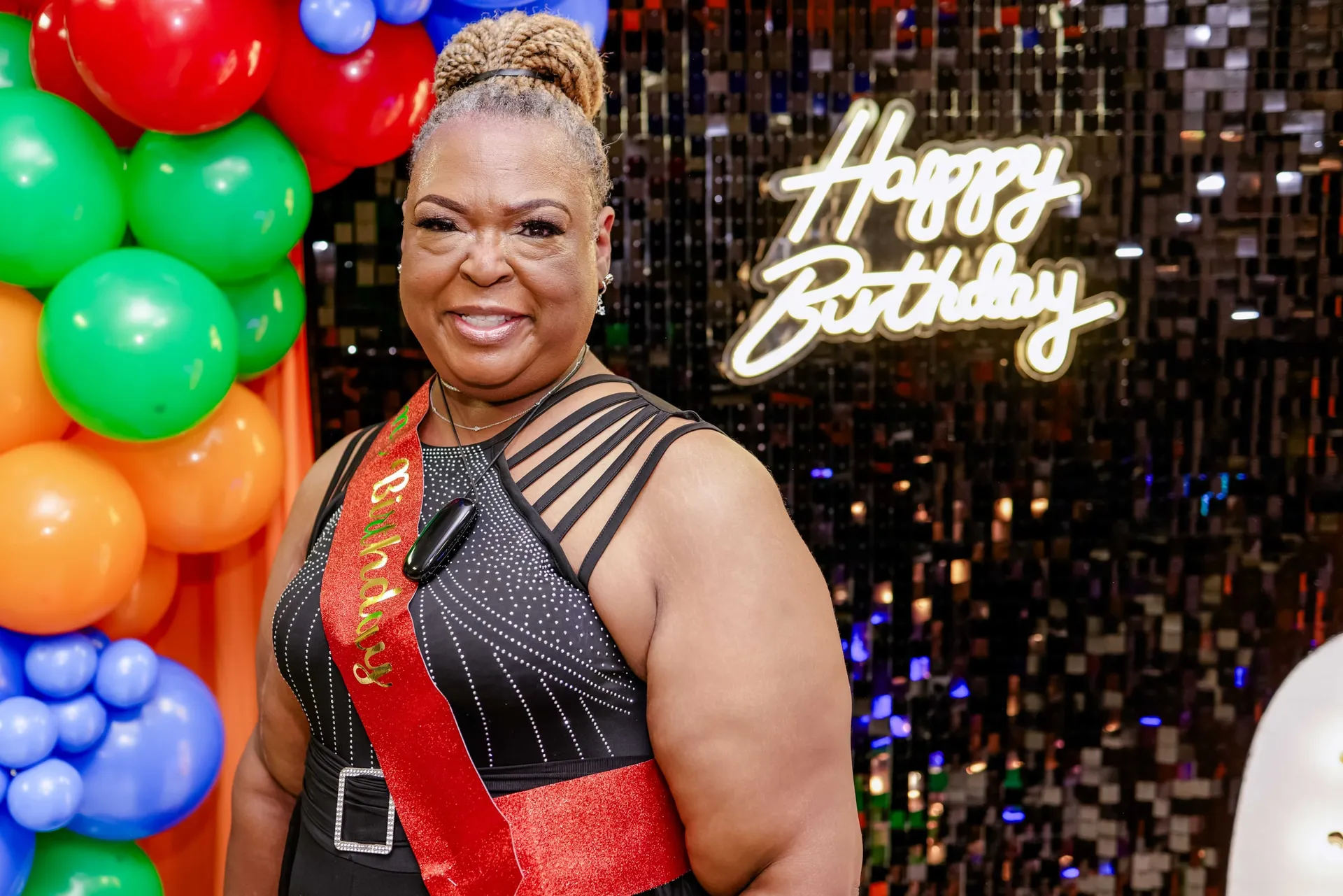 The birthday honoree in her sash before a neon Happy Birthday sign and balloon garland.