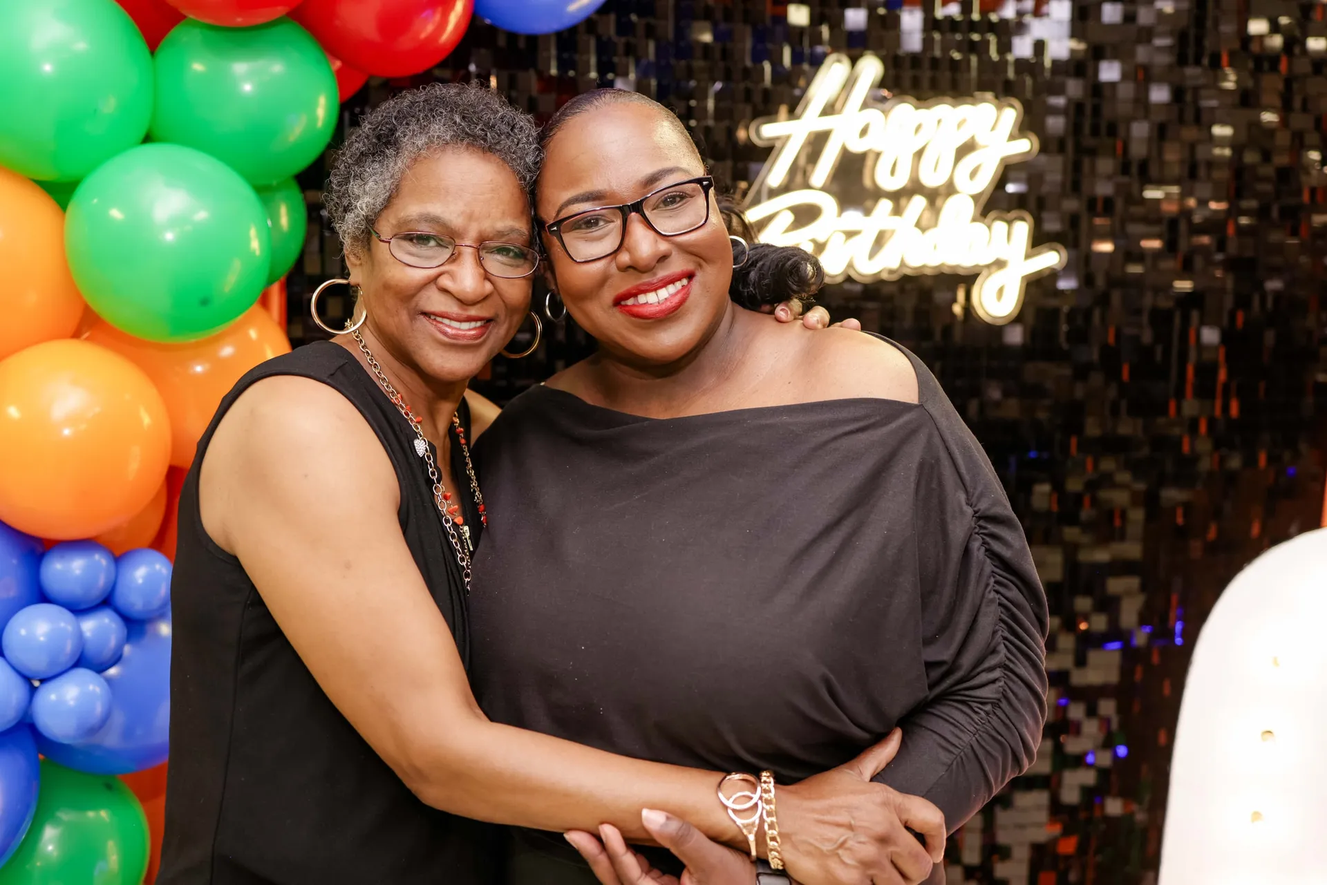 Two friends in a tight hug before the balloon garland and neon sign at a 60th.