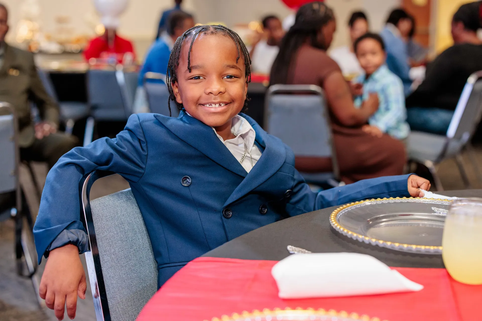 A young boy in a double-breasted blue suit smiling at his table during a banquet.