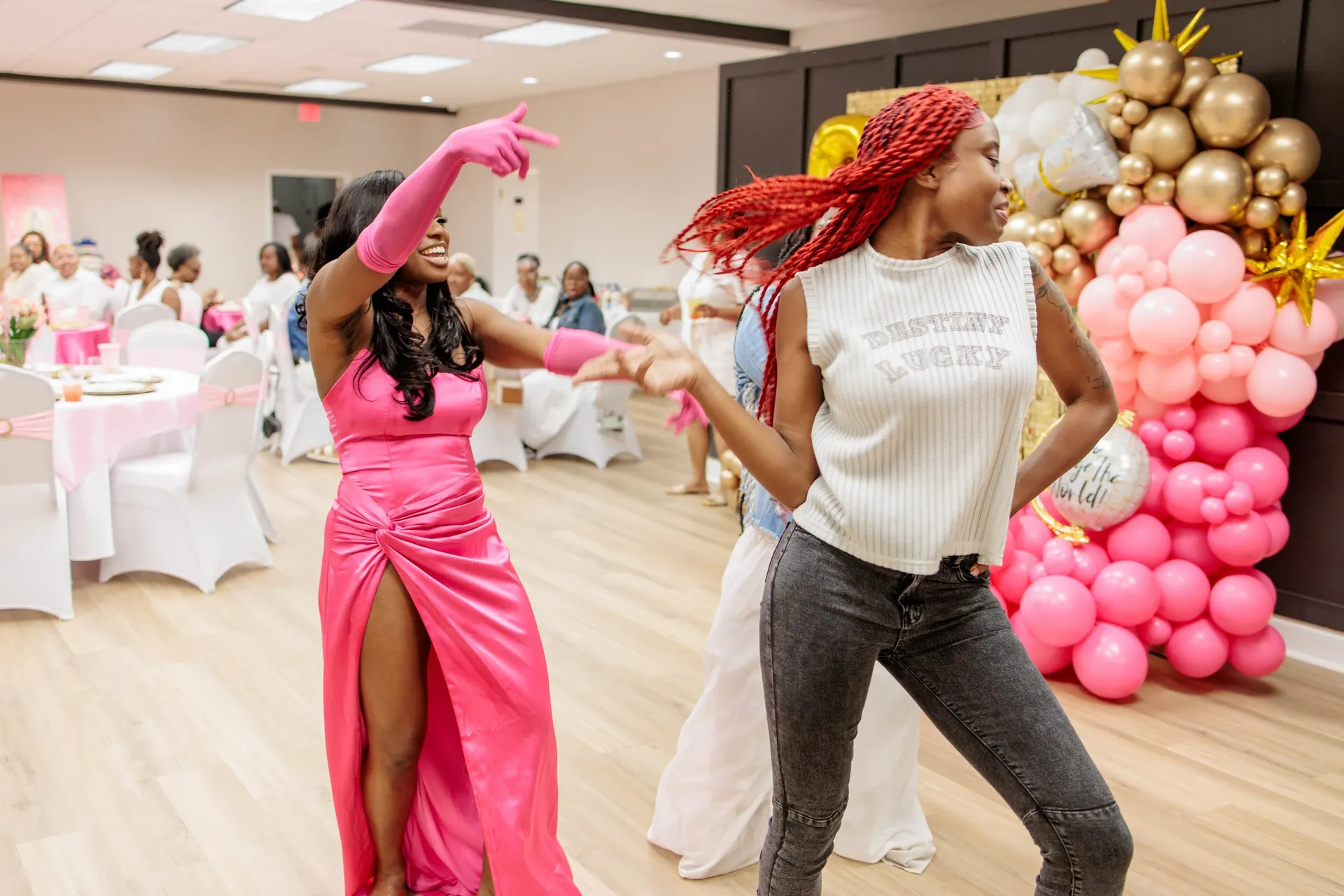 Two guests dancing in a hall of pink and gold balloons, one in pink satin.