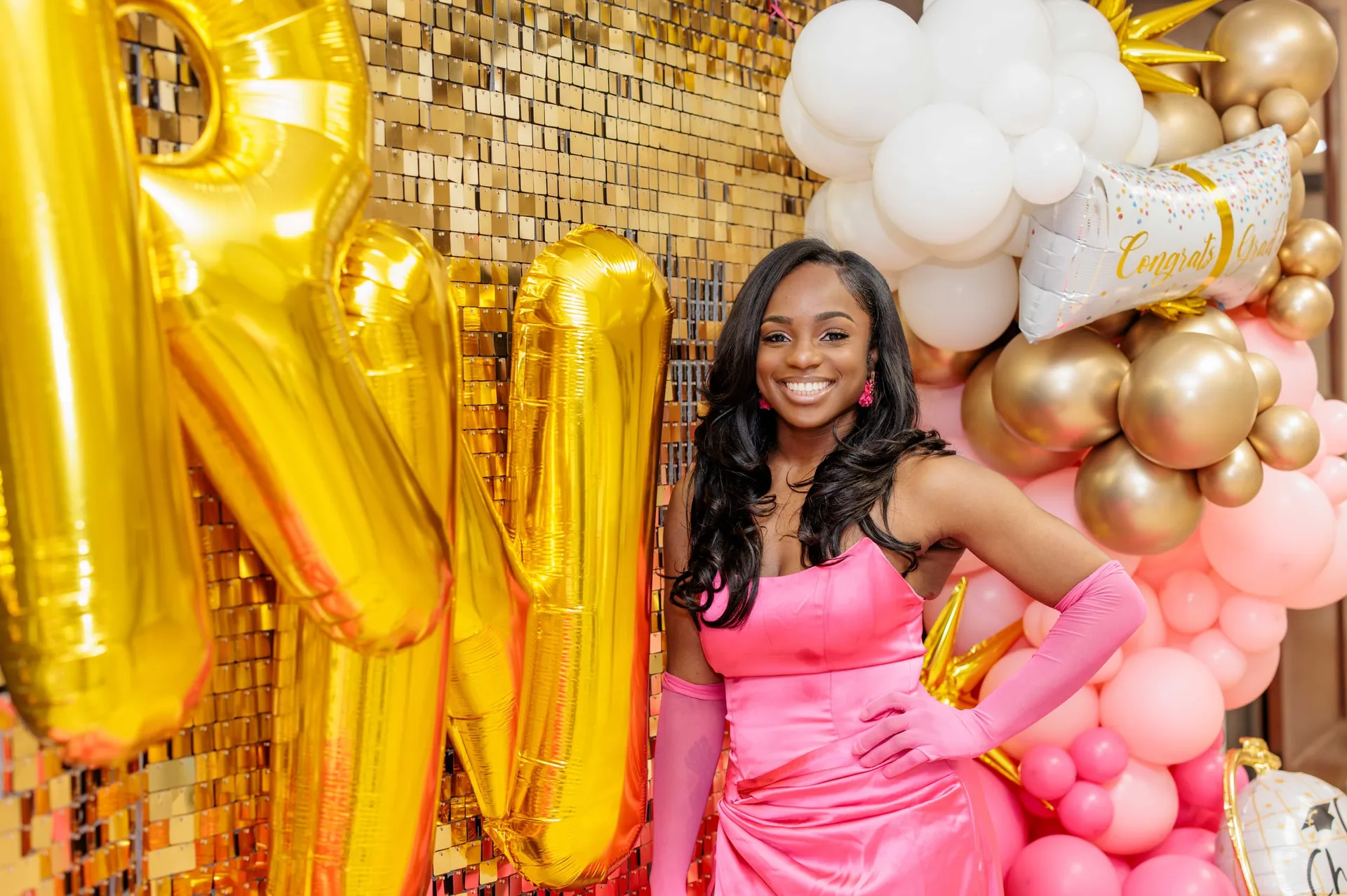 The guest of honor in a pink gown and gloves before a gold sequin wall.