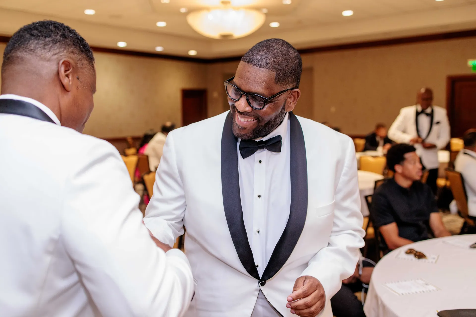 Two men in white dinner jackets laughing as they greet each other in a ballroom.