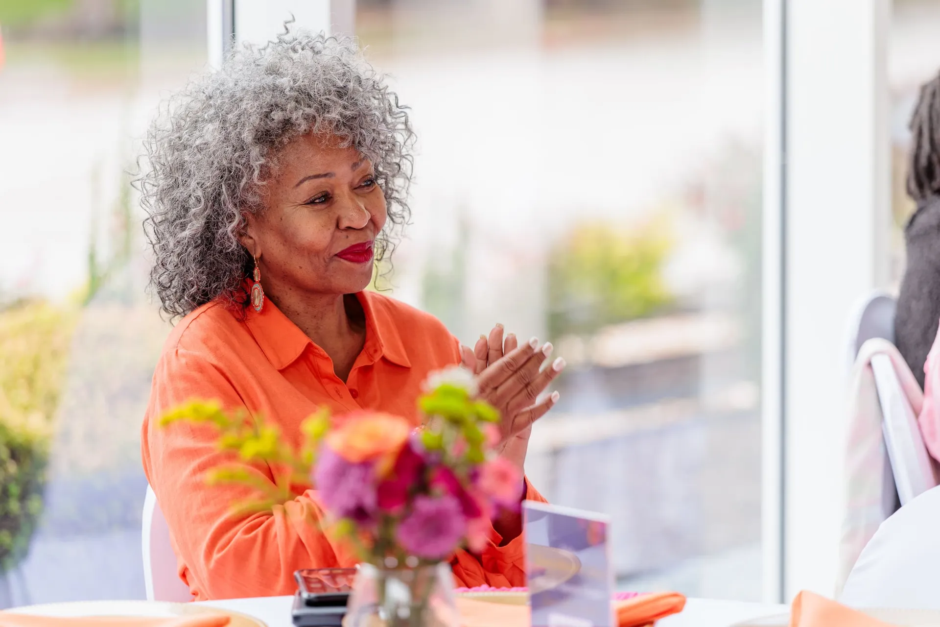 A guest in orange applauding from her table beside a bright bouquet at a summer soirée.