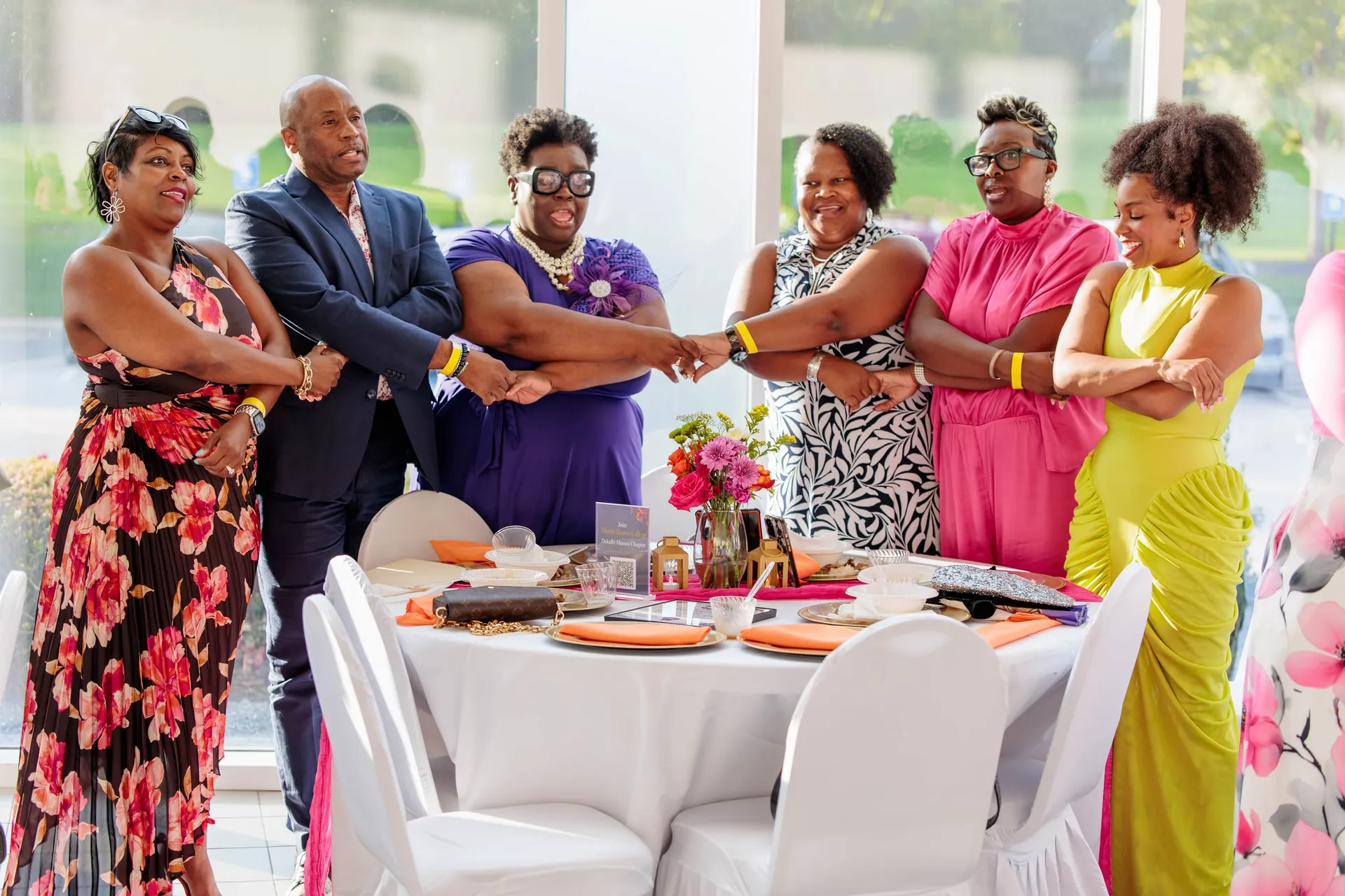 Six friends in bright summer dresses joining hands around a table at a soirée.