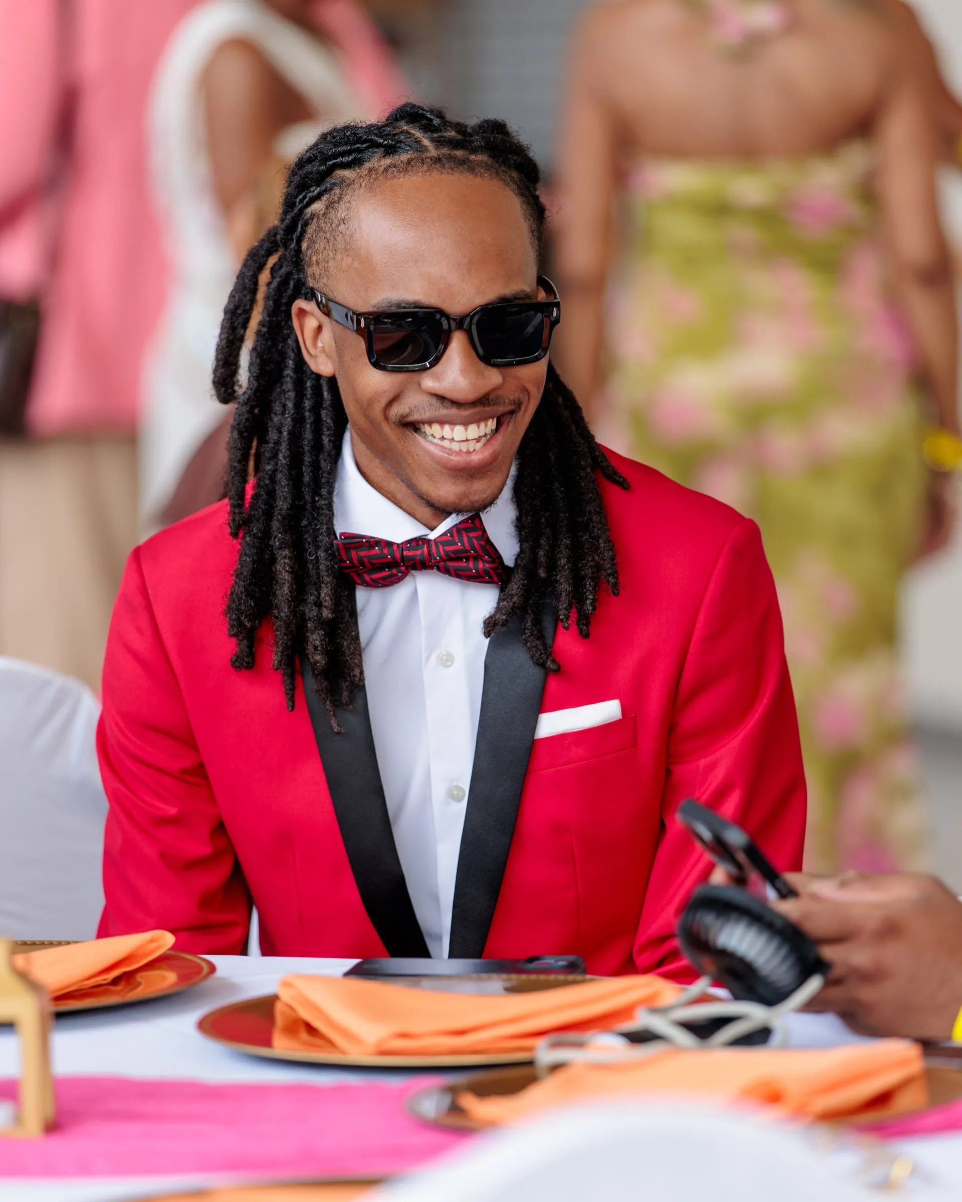 A guest in a red tuxedo jacket and dark glasses grinning at his soirée table.