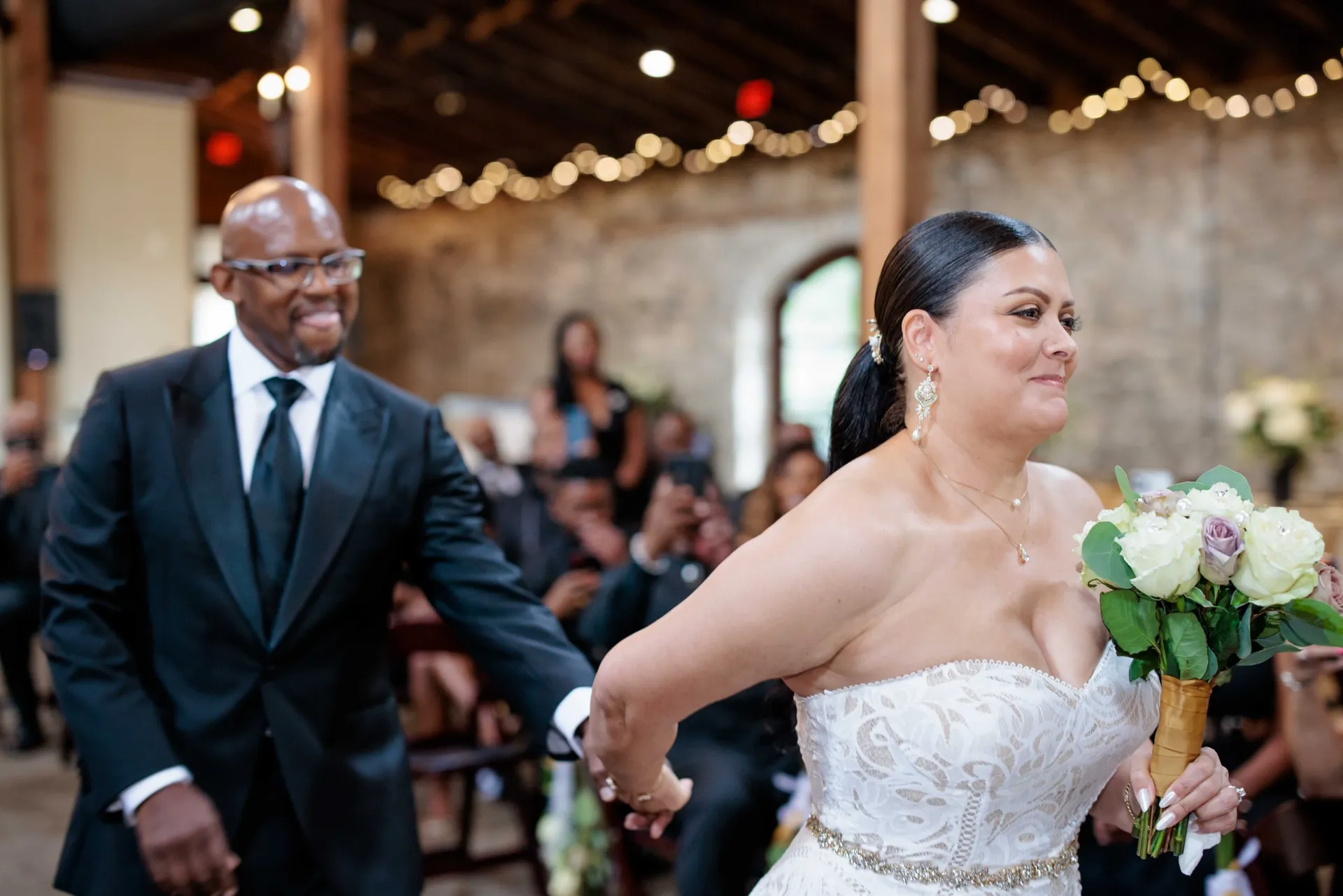 A bride in lace carrying a bouquet down the aisle under string lights.