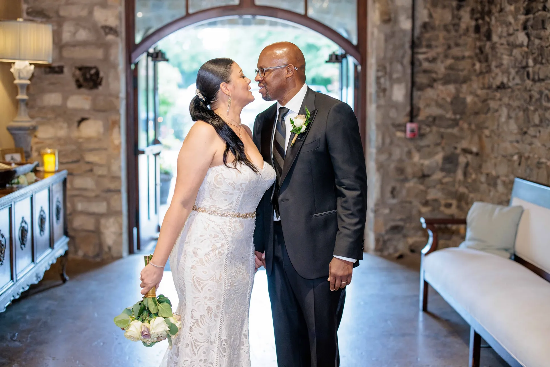 A couple nose to nose beneath a stone archway after renewing their vows, bouquet in hand.