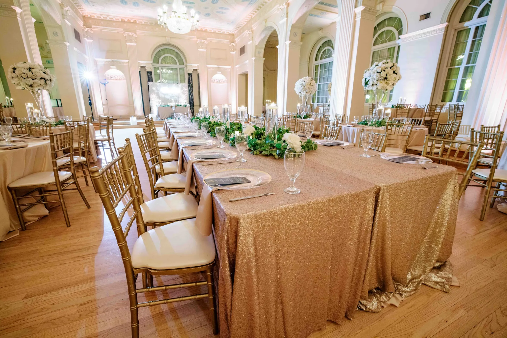 A long table in gold sequin linen and gold chairs in a white columned ballroom.