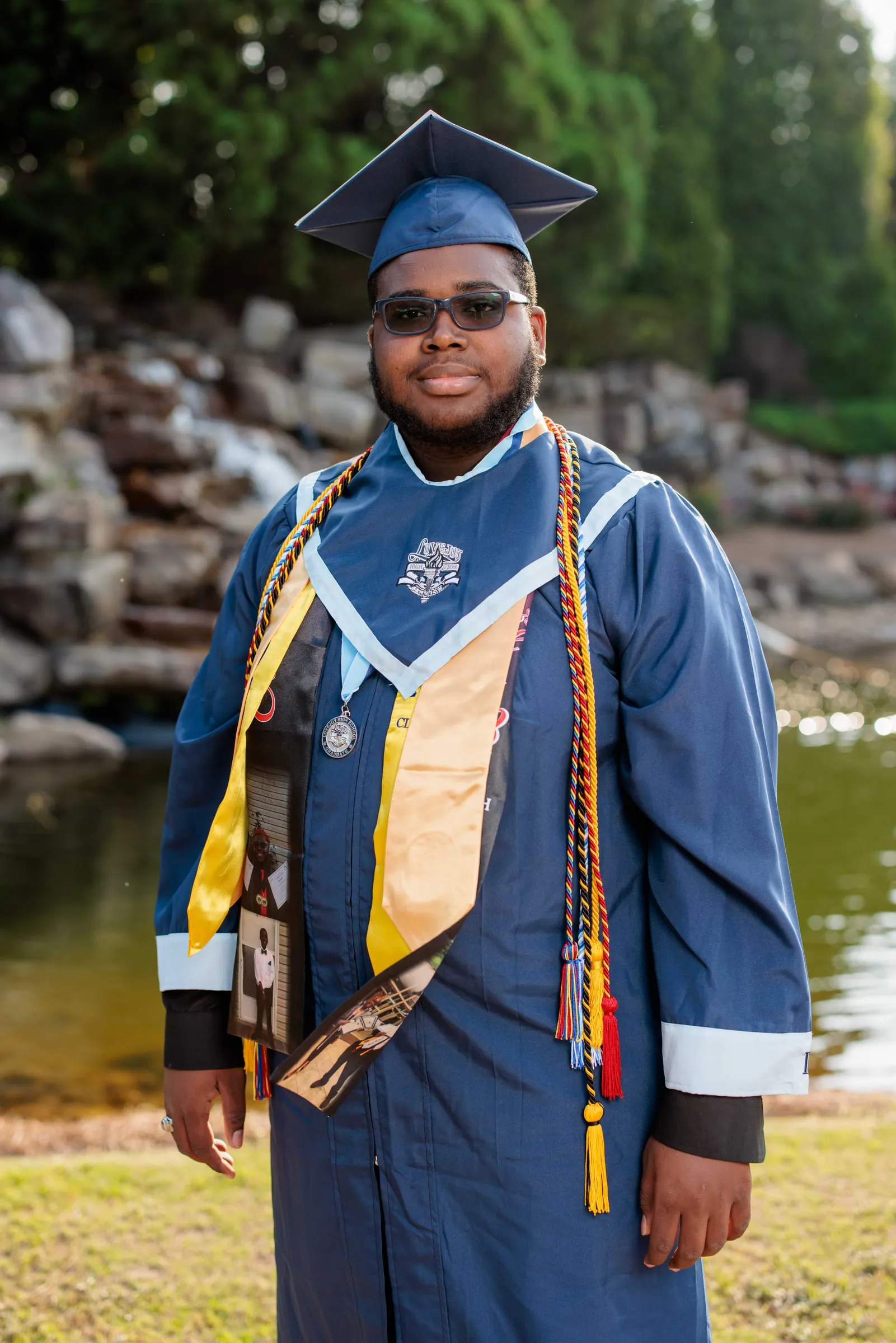 A graduate in a royal blue gown with gold honor cords standing before a rocky waterfall.