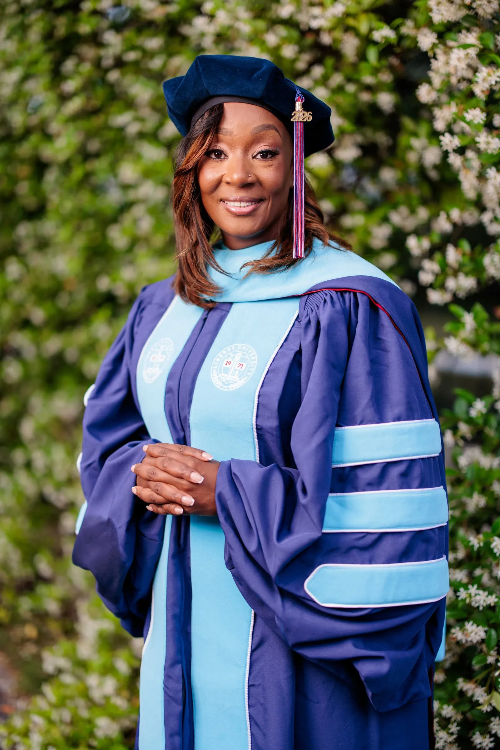 A doctoral graduate in navy and sky blue regalia with a velvet tam before white jasmine.