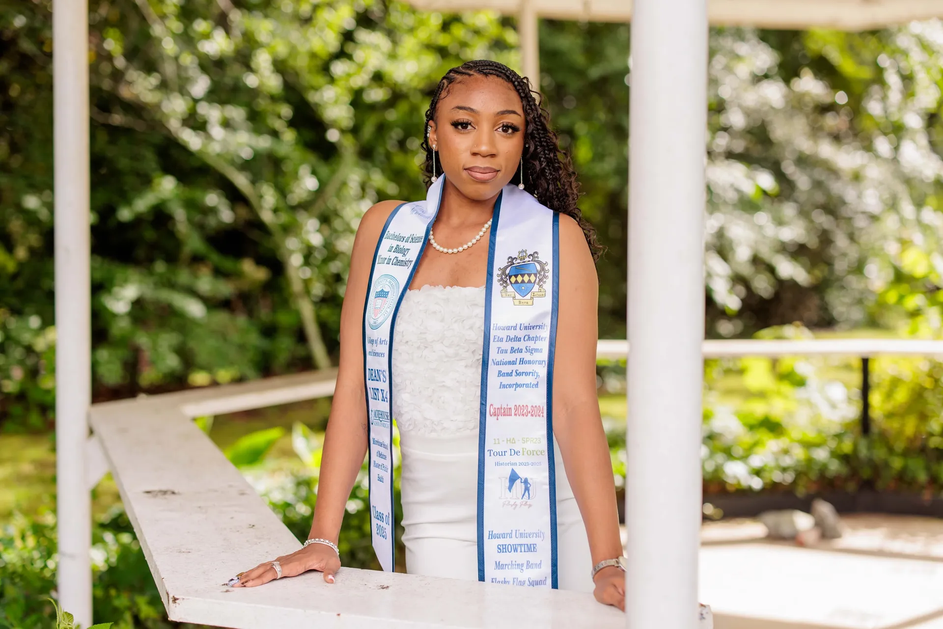 A graduate in white with an honor stole standing at the rail of a garden gazebo.