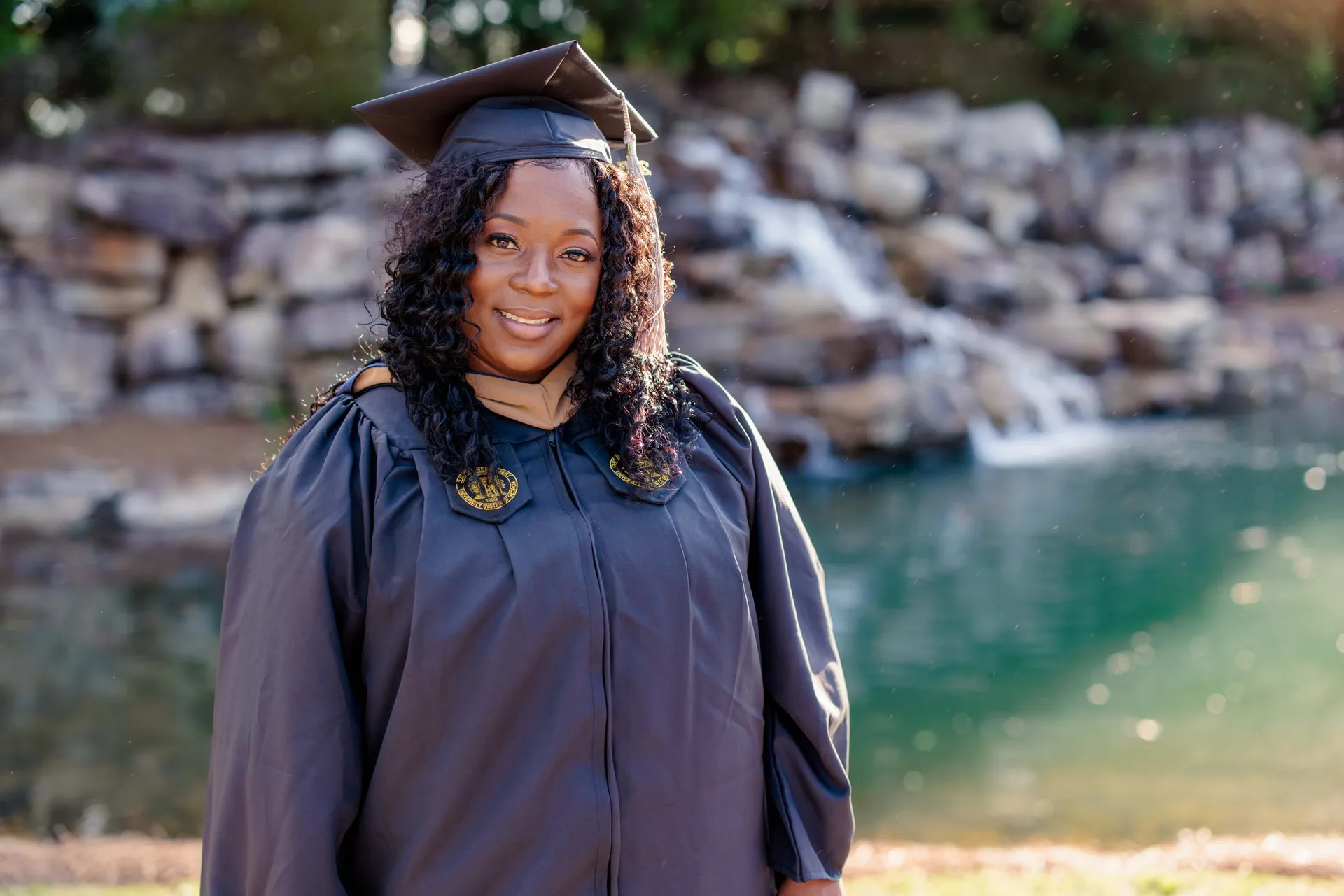 A graduate in her navy cap and gown smiling before a rocky waterfall and green pool.