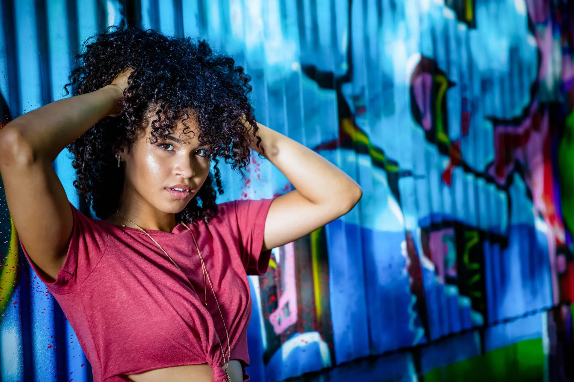 A senior in a red tee, hands in her curls, against a blue graffiti wall.