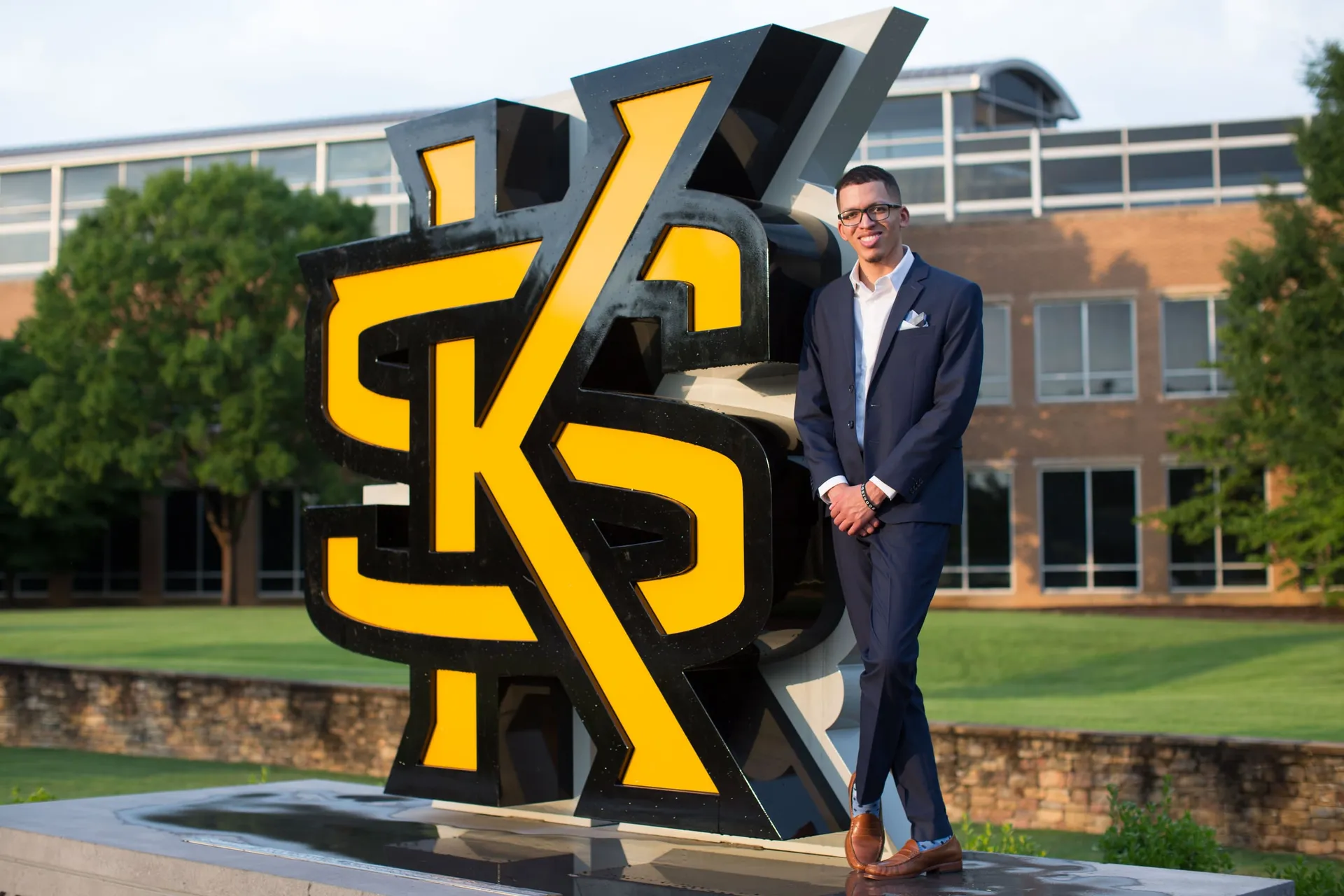 A graduate in a navy suit beside the giant black and gold KS letters on campus.