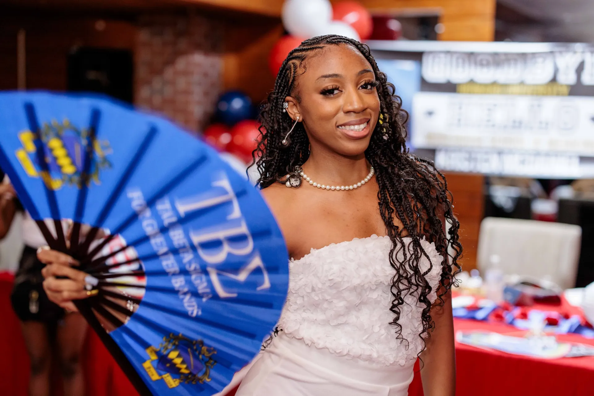 A graduate in white holding a large blue band-sorority fan at her graduation party.