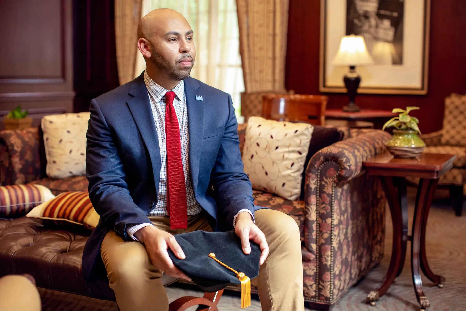 A graduate in a blue suit and red tie in a leather armchair, tam in hand.
