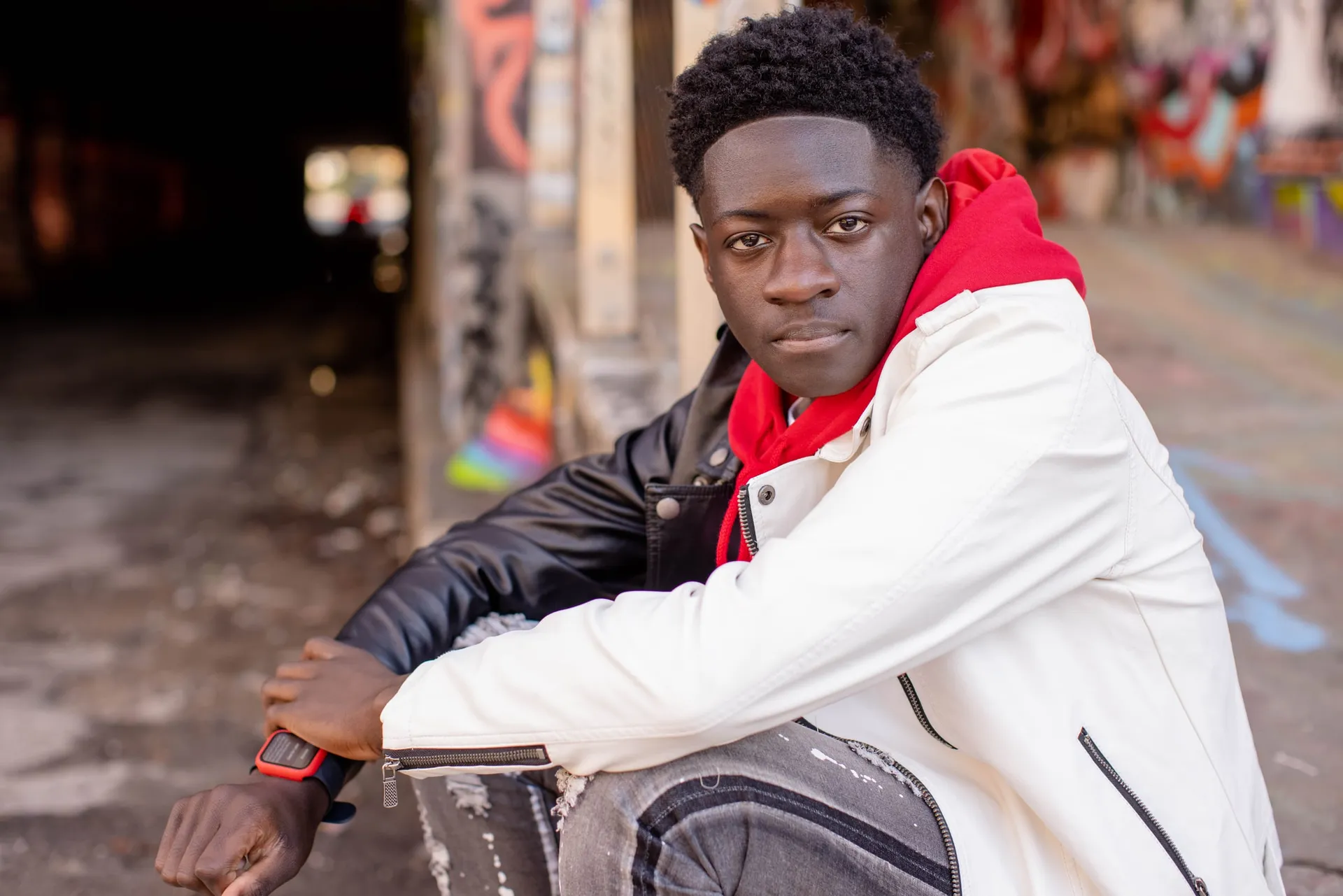 A young man in a white and red jacket seated in a graffiti-covered tunnel.