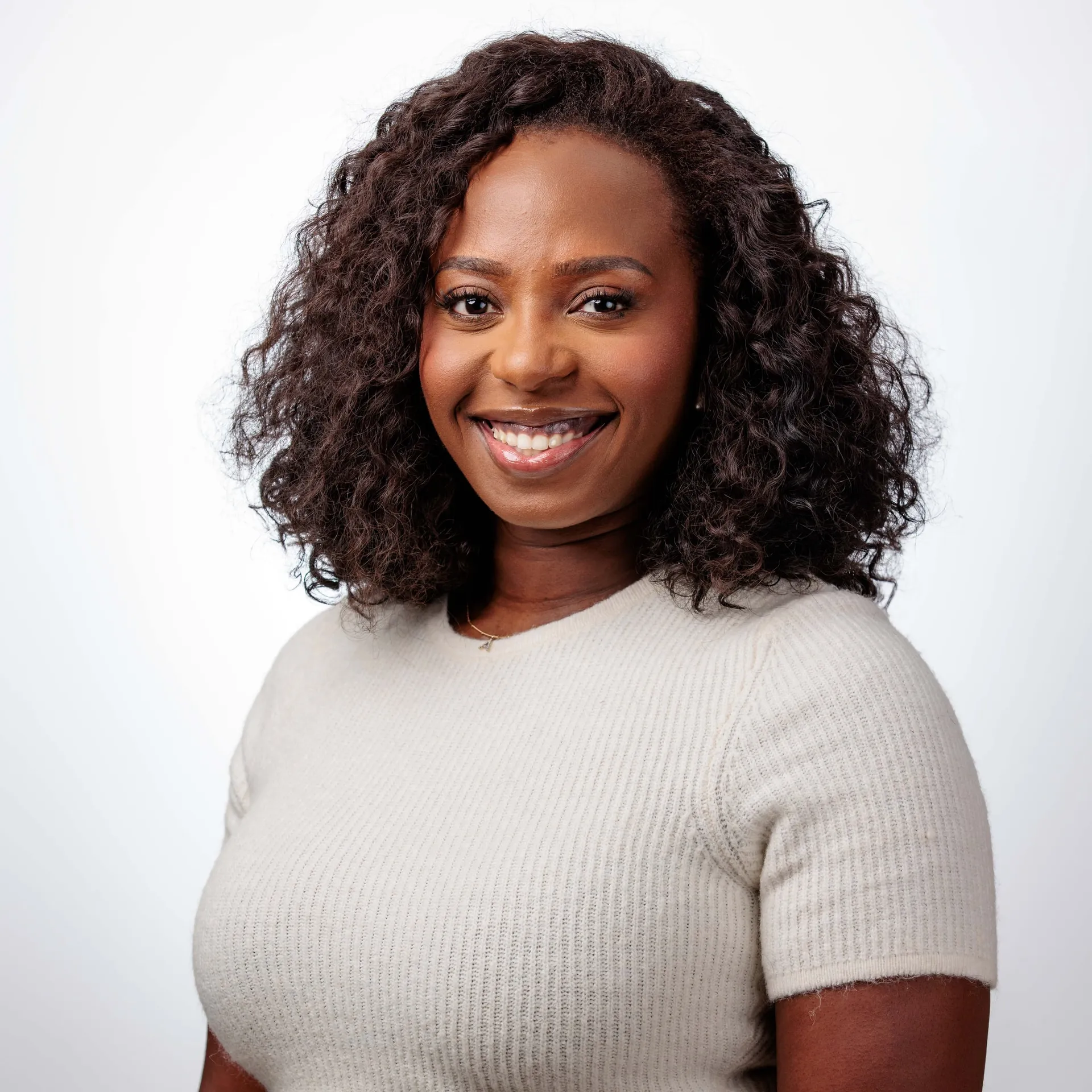 A studio headshot of a woman with long curls in a beige knit top on white.