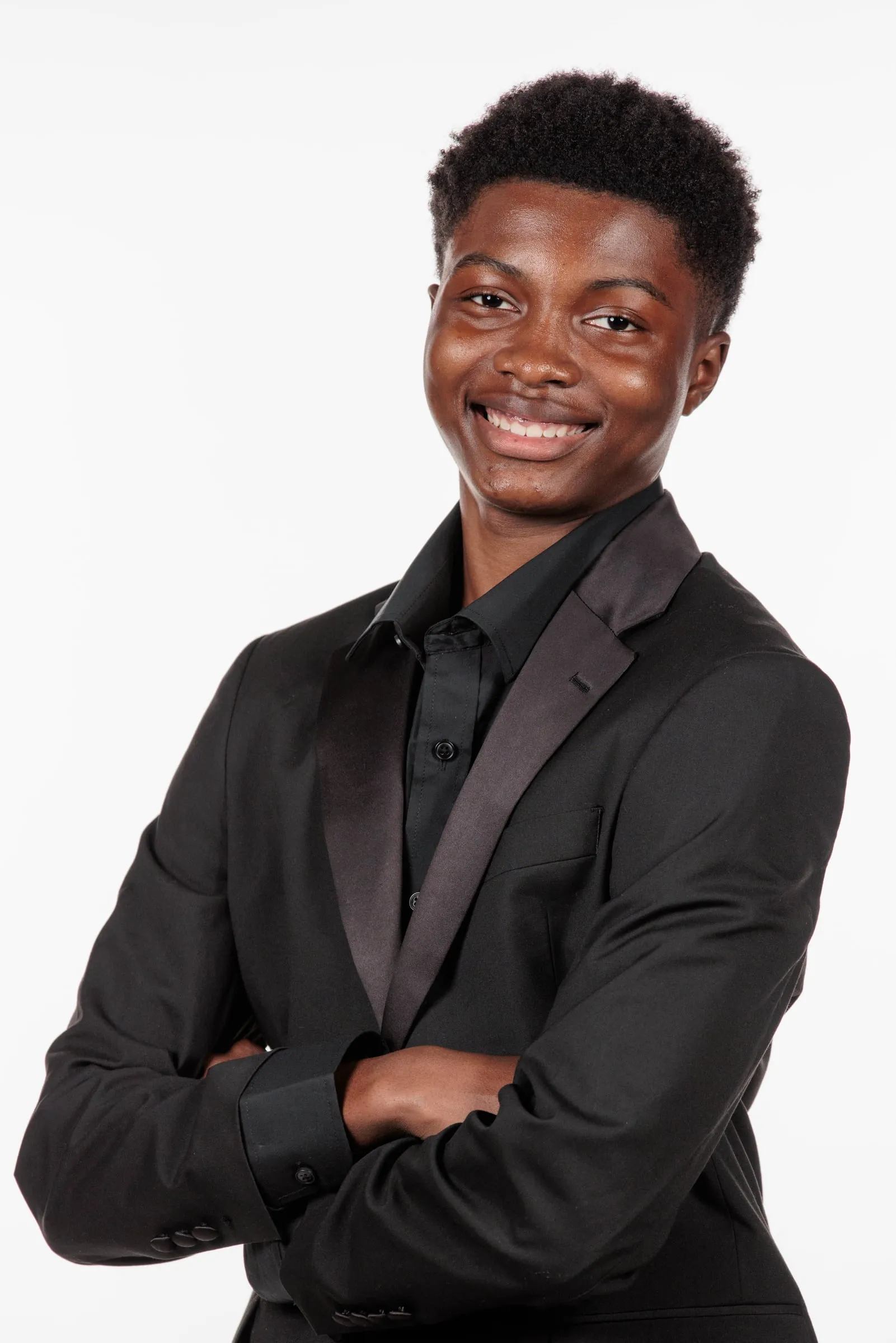 A young man in a black suit with arms crossed smiling in a white studio headshot.