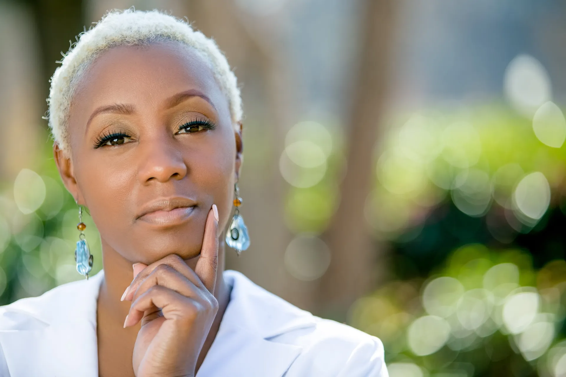 A woman with cropped blonde hair in a white blazer, chin on hand, against green bokeh.