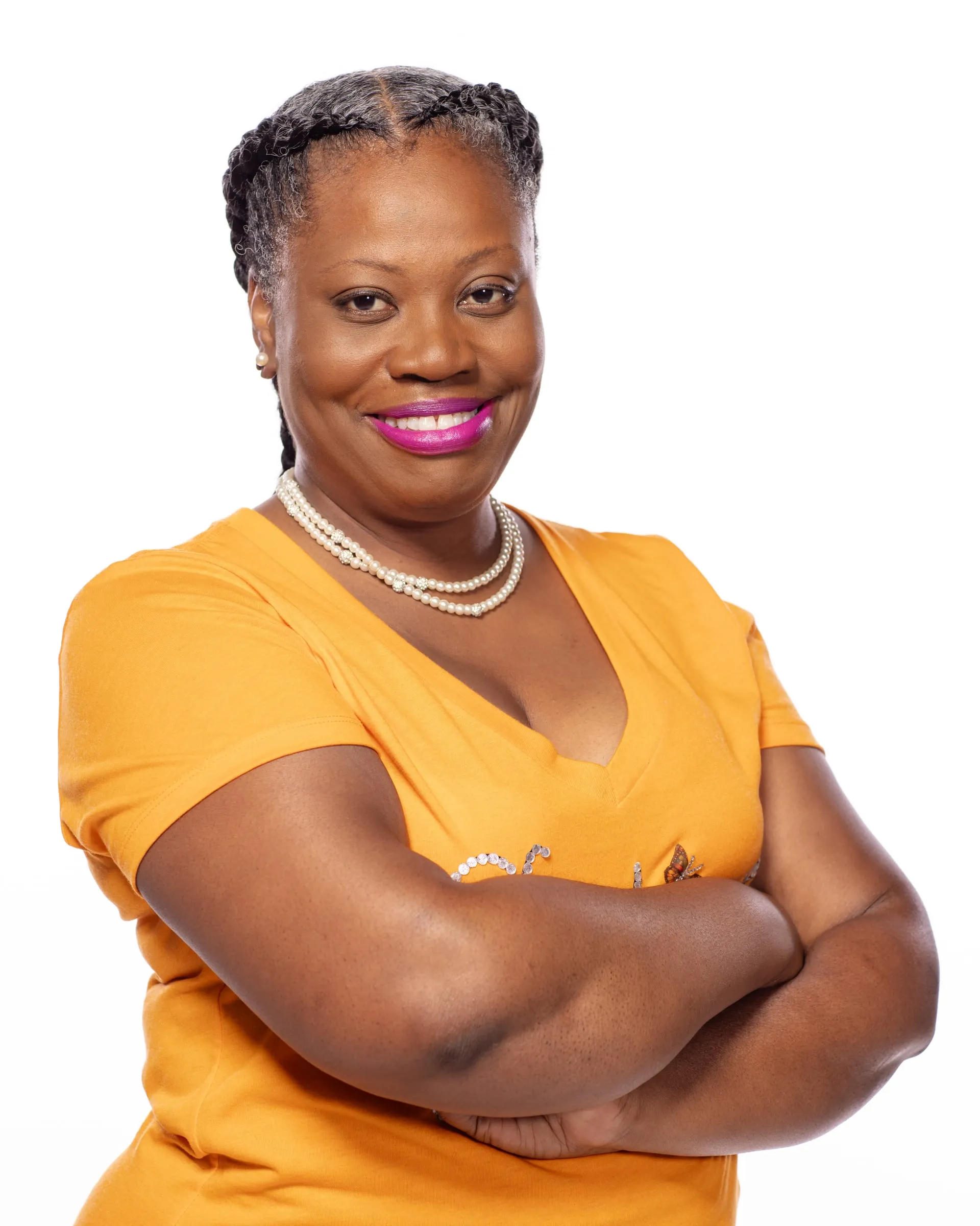 A studio headshot of a woman in a mustard top and pearls, arms crossed, smiling.