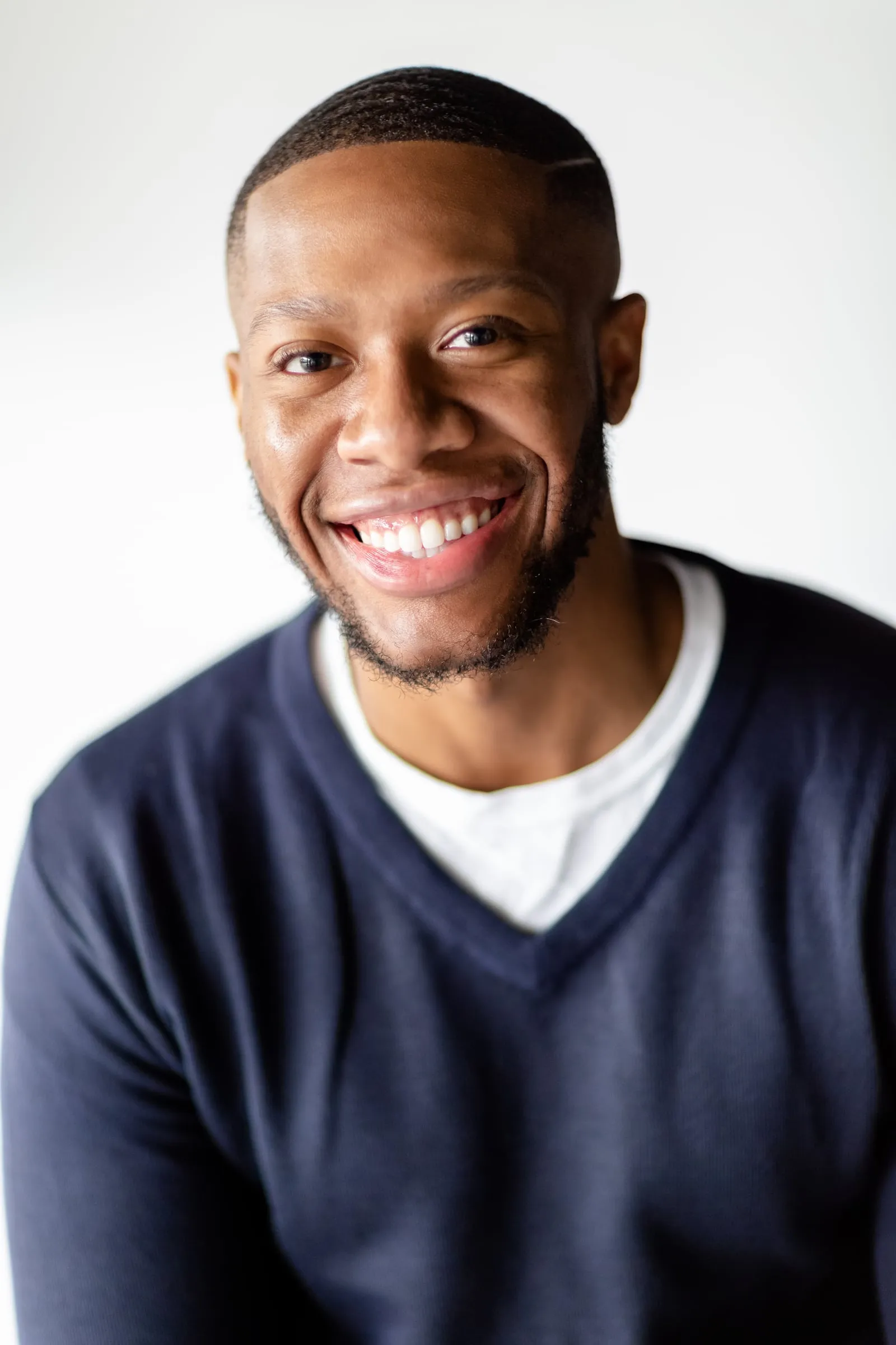 A man in a navy v-neck sweater over a white tee laughing on grey.