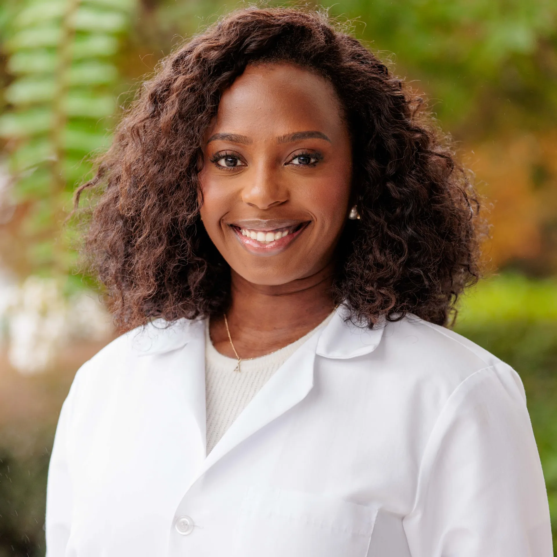 A physician in a white coat smiling warmly before soft green foliage.