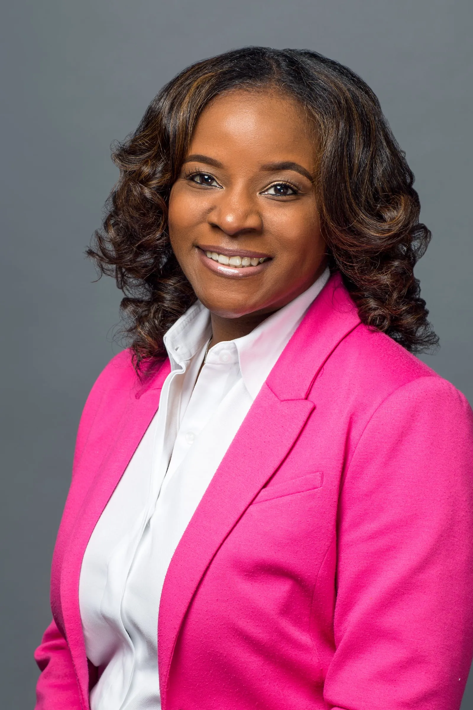 A studio headshot of a woman with curled hair in a hot pink blazer on grey.