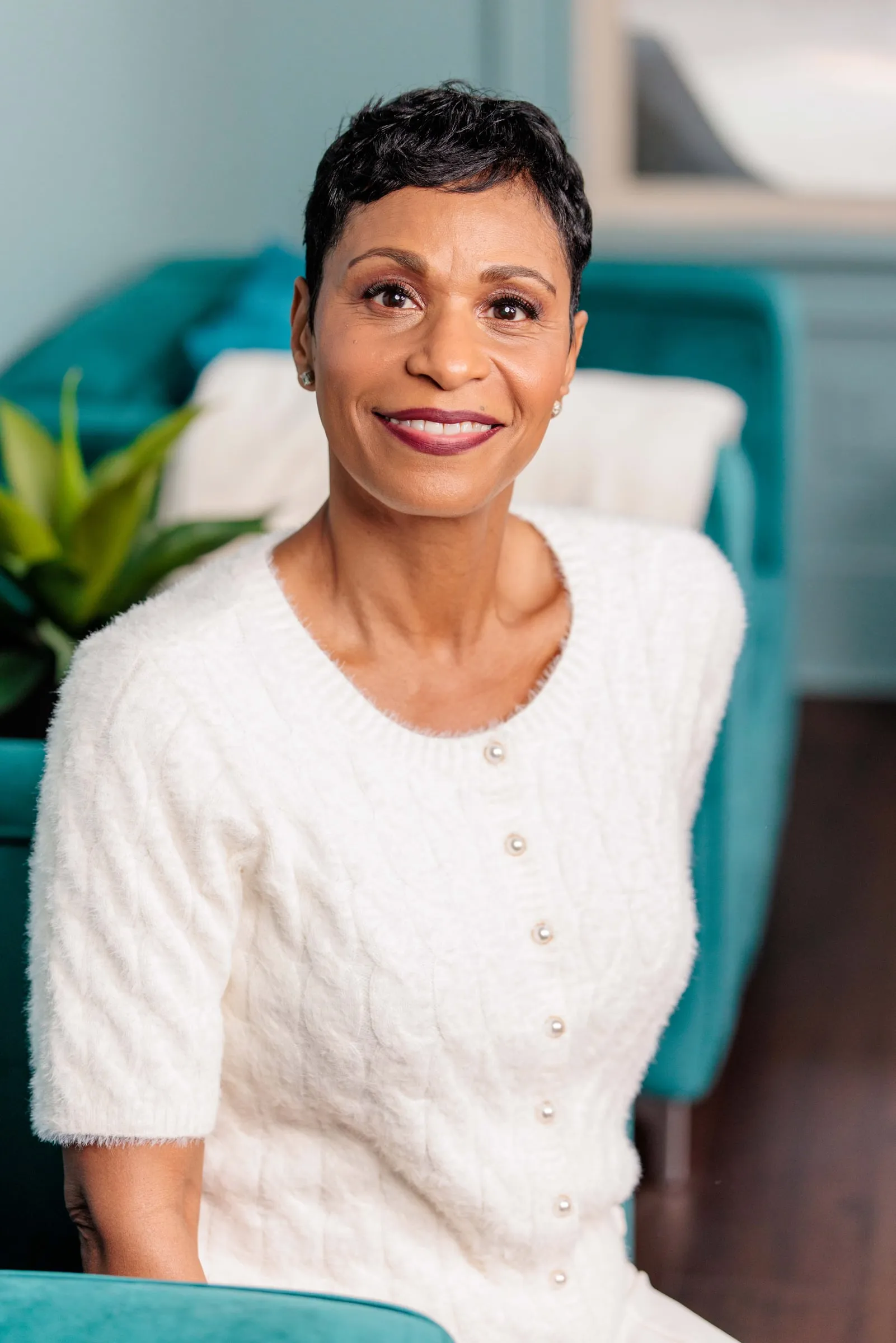 A woman with cropped hair in a white sweater seated in a teal armchair, smiling.