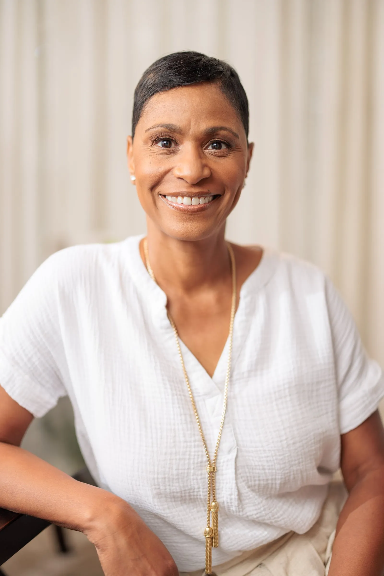 A woman with cropped hair in a white blouse and long pendant in soft window light.