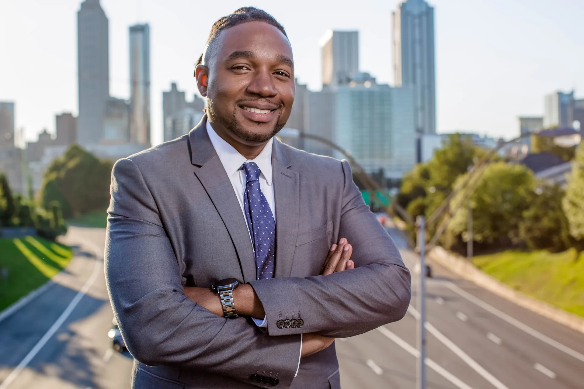A man in a grey suit, arms crossed, on a bridge above the Atlanta skyline highway.