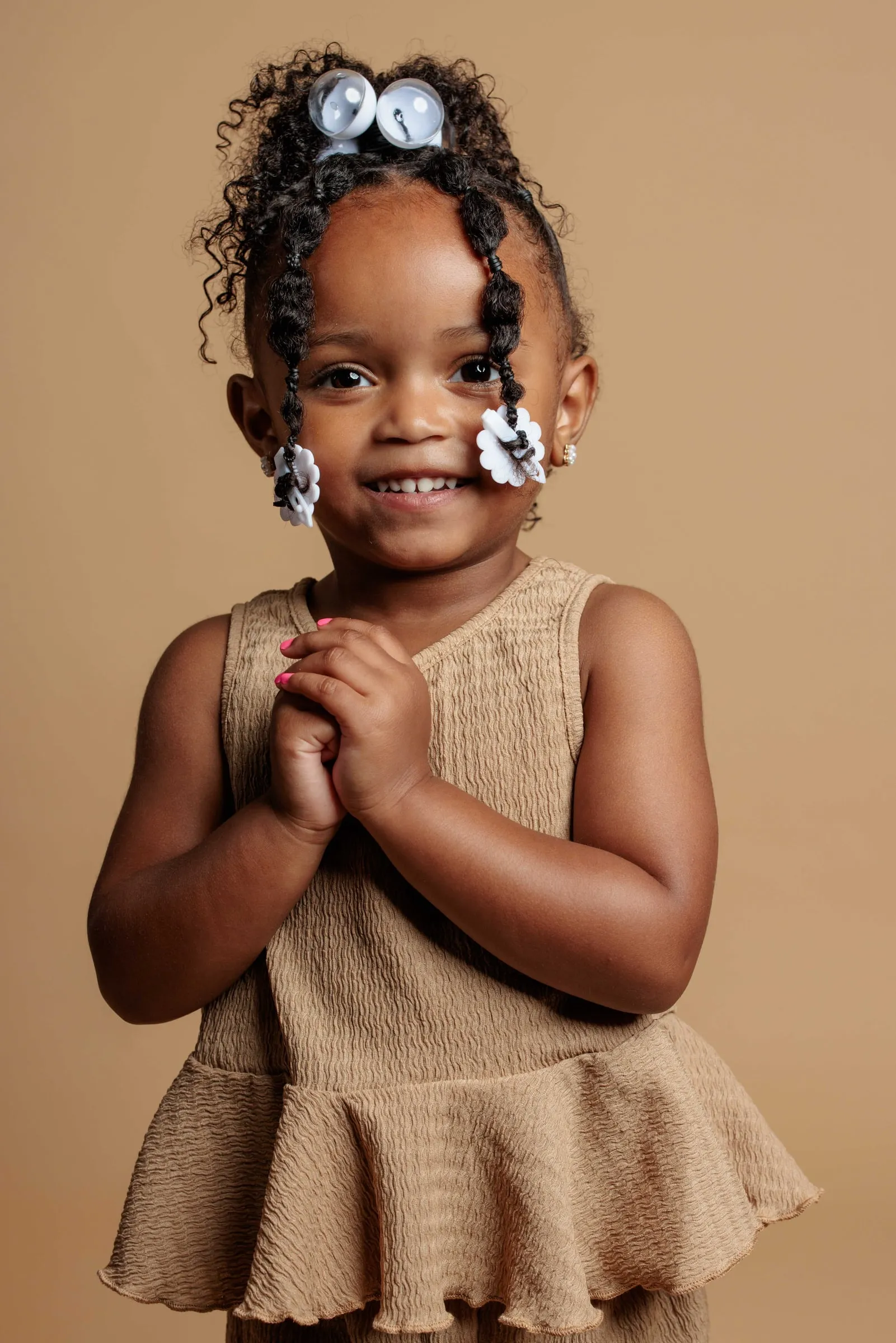 A little girl in a tan ruffled dress and hair bows clasping her hands and grinning.