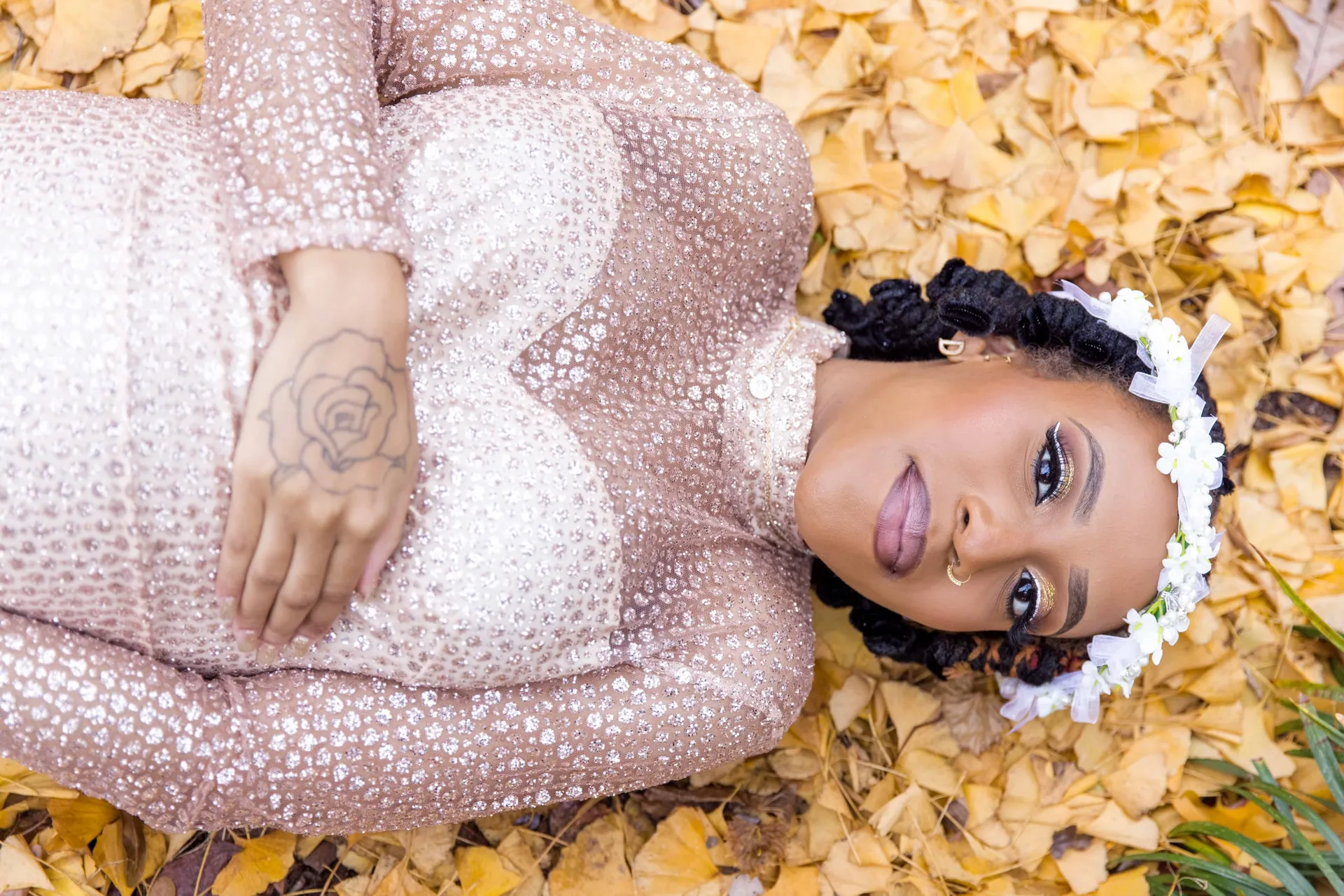 An expectant mother in sequins and a flower crown lying in a bed of yellow leaves.