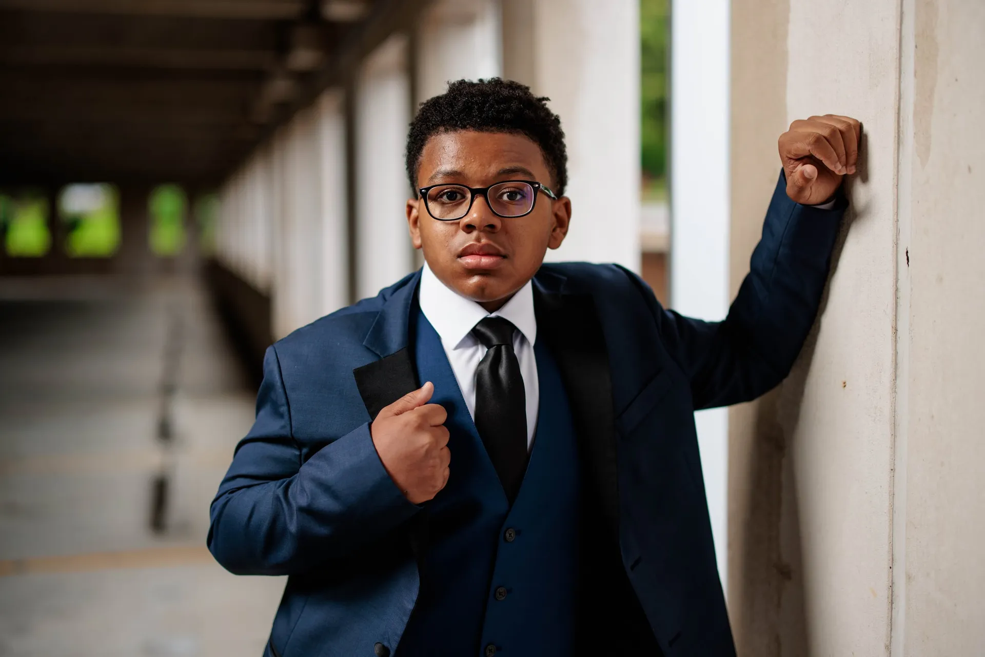A boy in glasses and a navy three-piece suit posing against a pillar in a colonnade.