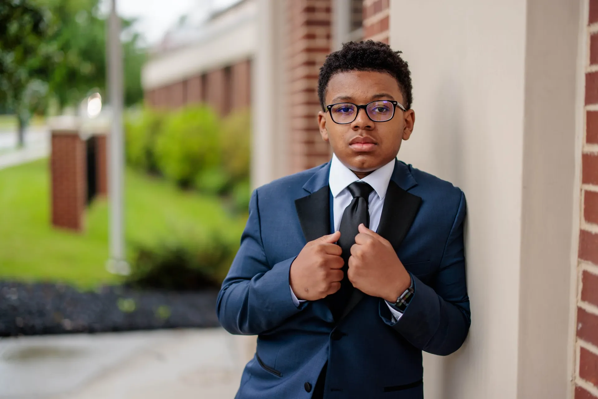 A boy in a navy suit adjusting his lapels beside a brick wall on a lawn.